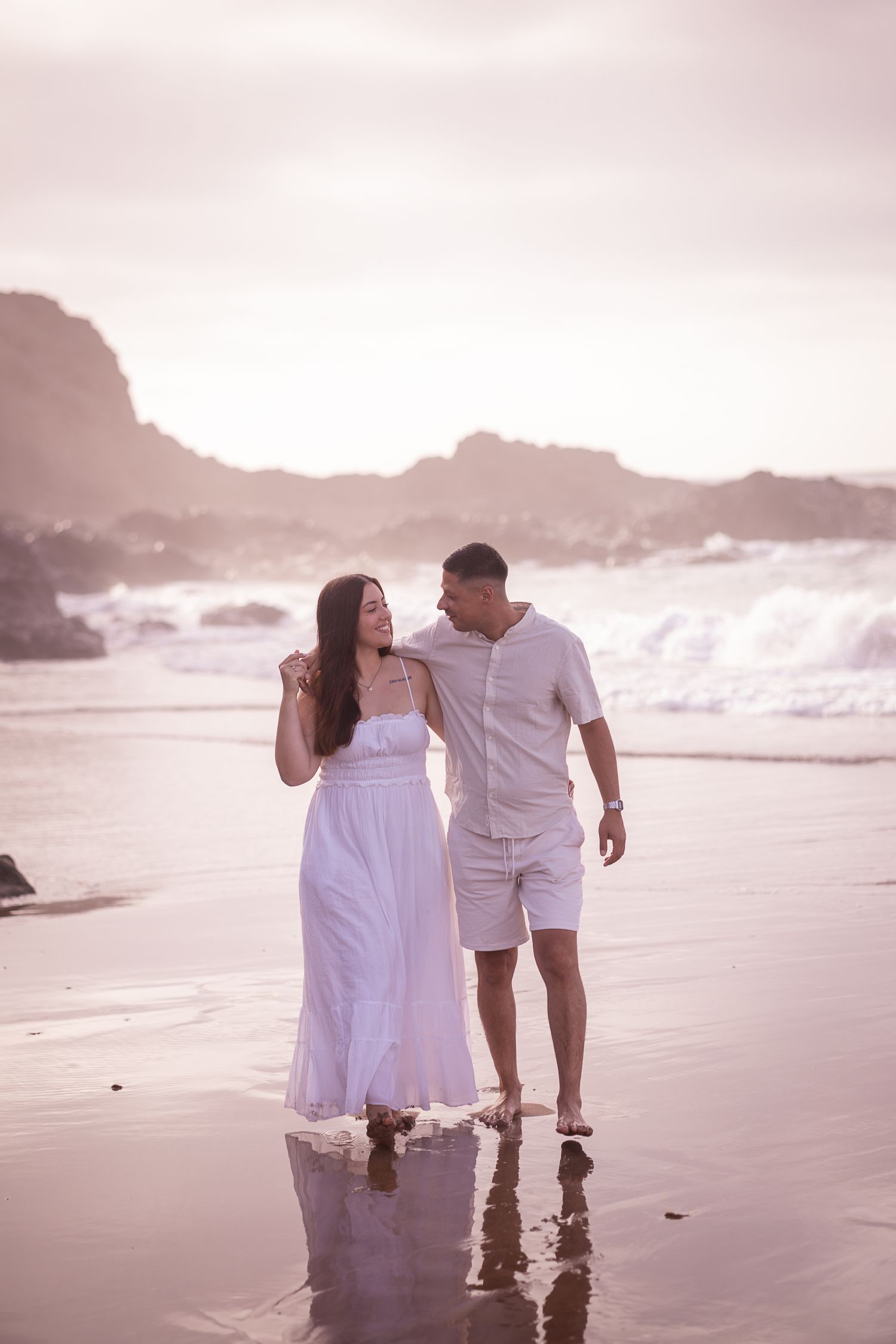 Romantic beach photoshoot with couple walking along misty shoreline in white outfits.