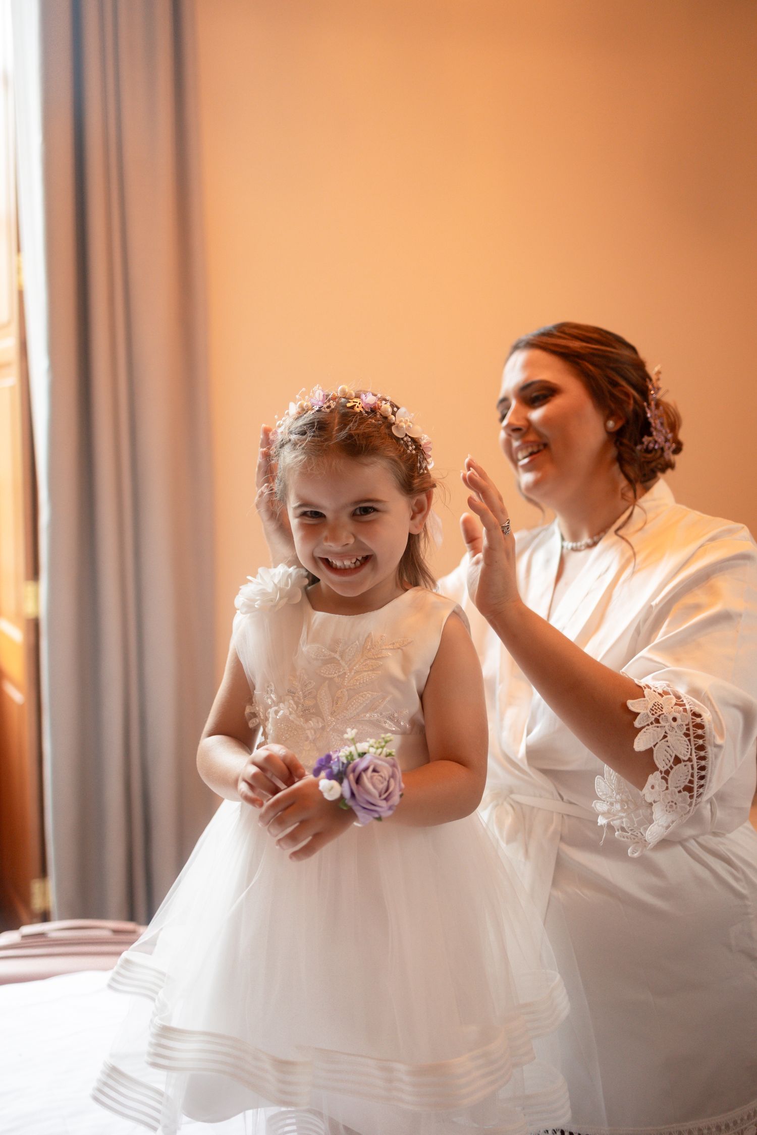 Smiling bride in white robe adjusting flower girl hair accessory, both wearing white dresses with purple floral accents.