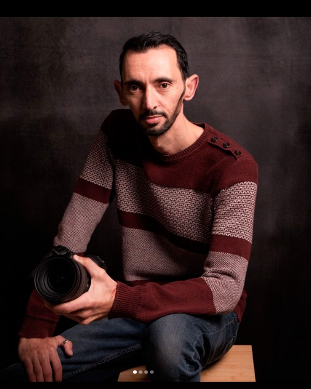 Professional photographer wearing a burgundy and gray patterned sweater holds a camera lens in a studio portrait.
