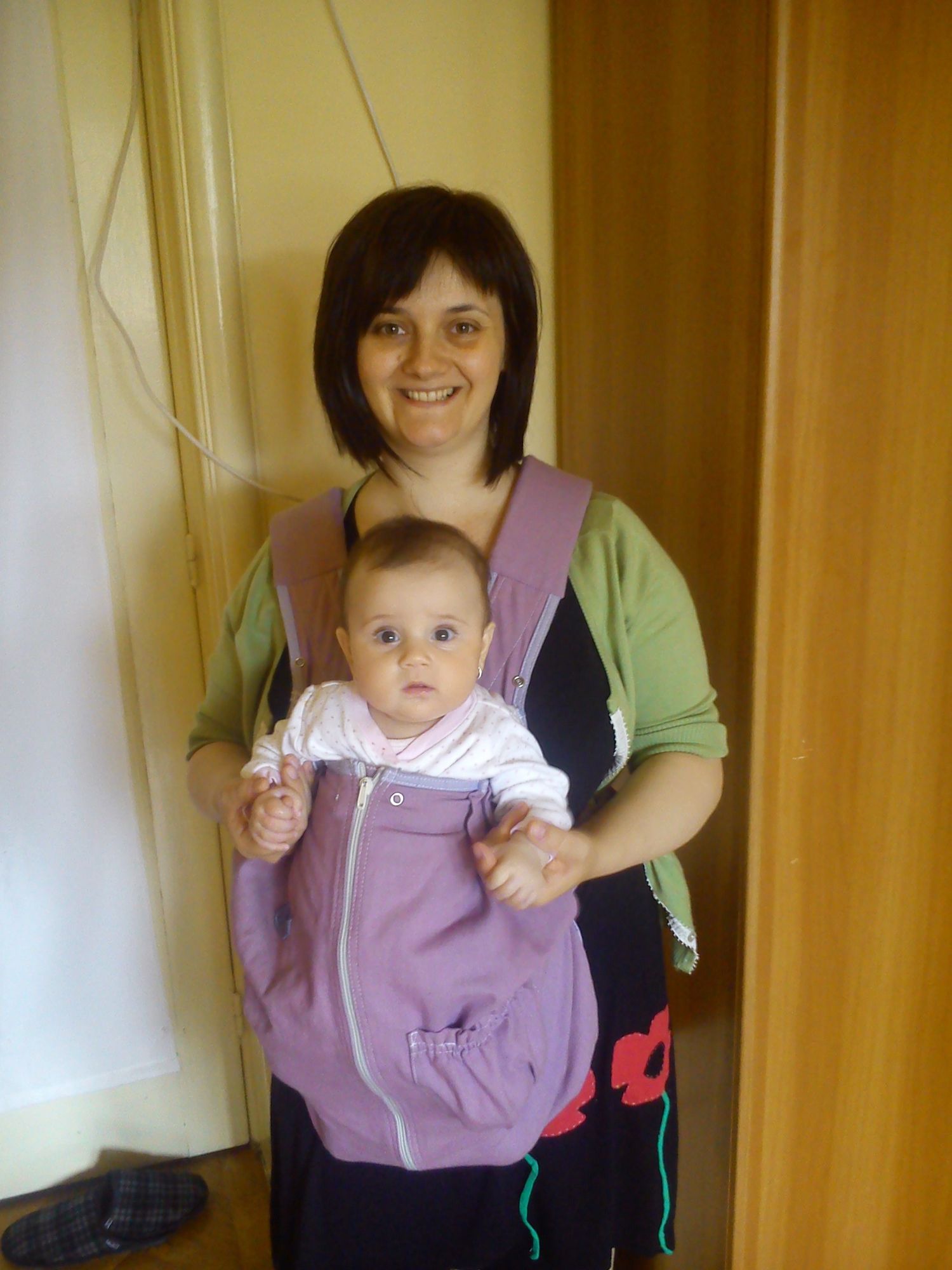 A baby wearing purple overalls being carried in a baby carrier.