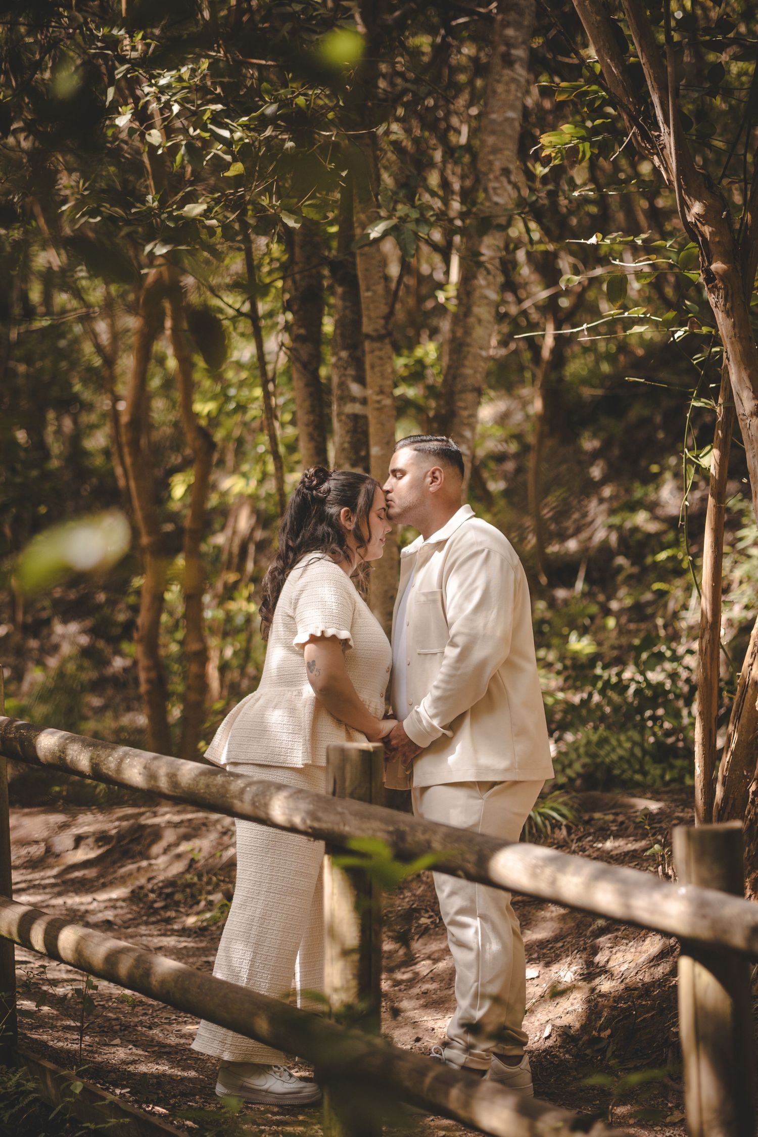 Couple sharing a romantic kiss on a wooden path surrounded by lush green forest trees in natural light.