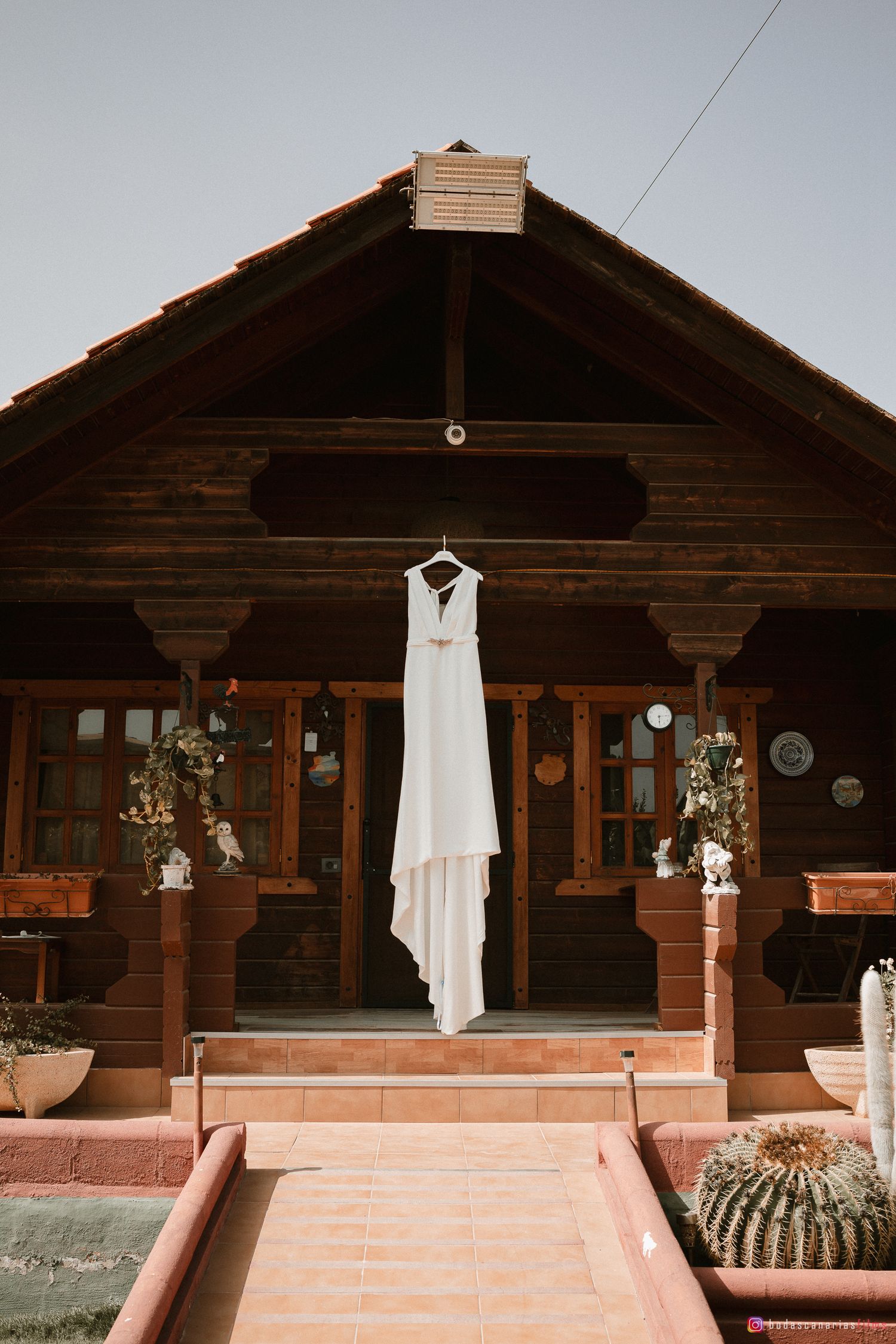 A white wedding dress hangs outside a rustic wooden cabin with terracotta steps.