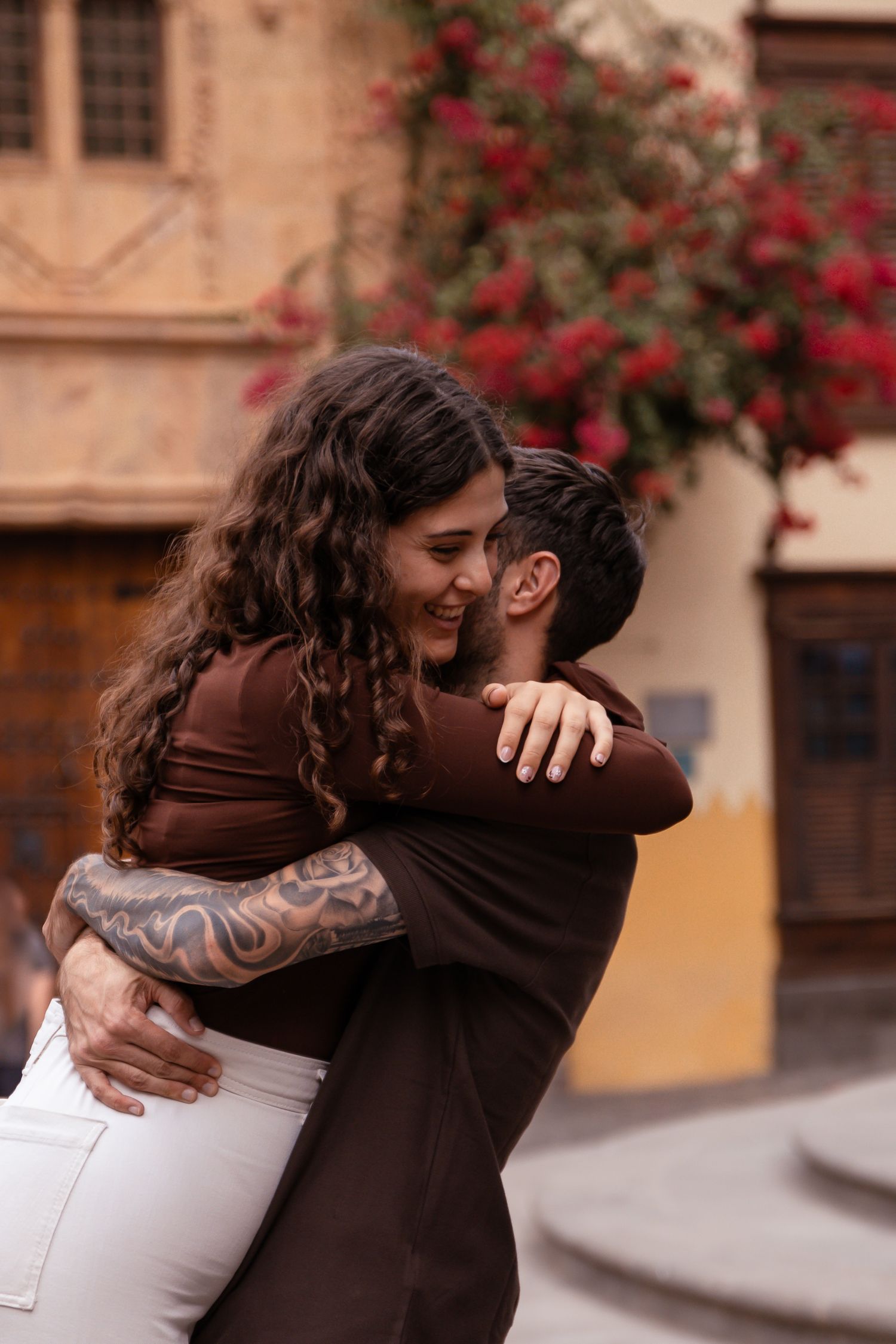 Couple sharing an intimate embrace in front of a building with blooming red flowers.