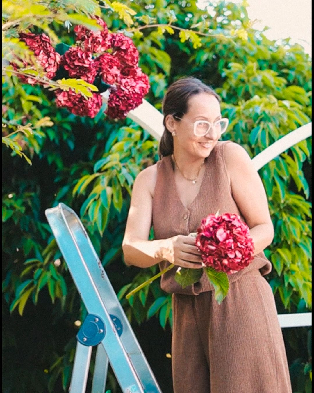 A person in a brown dress holds vibrant red flowers while standing near lush greenery and a blue ladder.