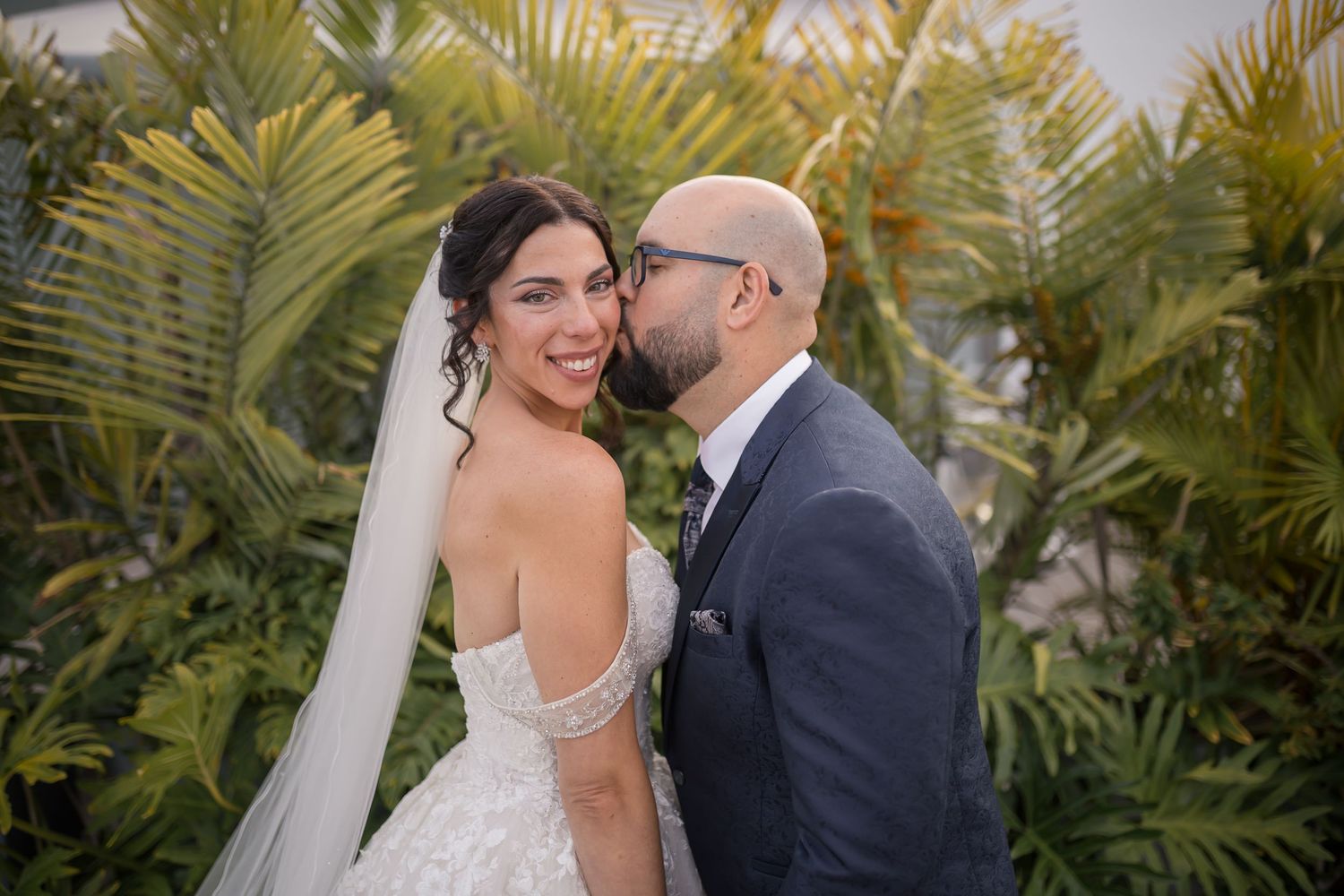 A couple shares an intimate moment on their wedding day surrounded by lush tropical palm fronds in an outdoor setting.