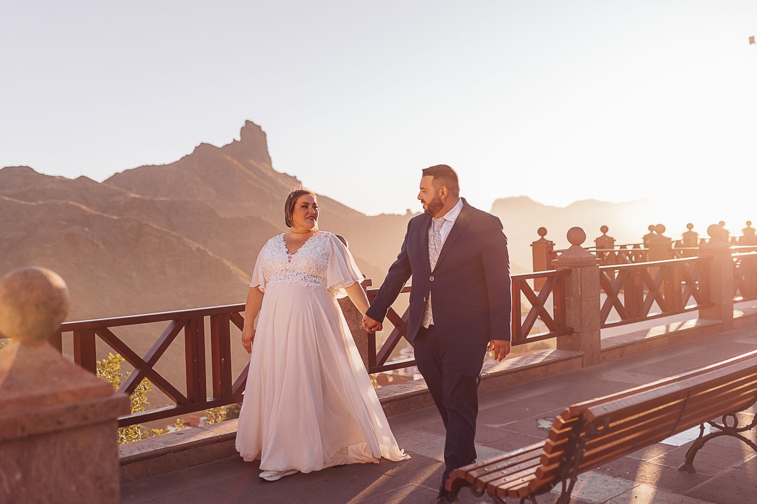 A couple in wedding attire walks along a scenic mountain path at sunset.