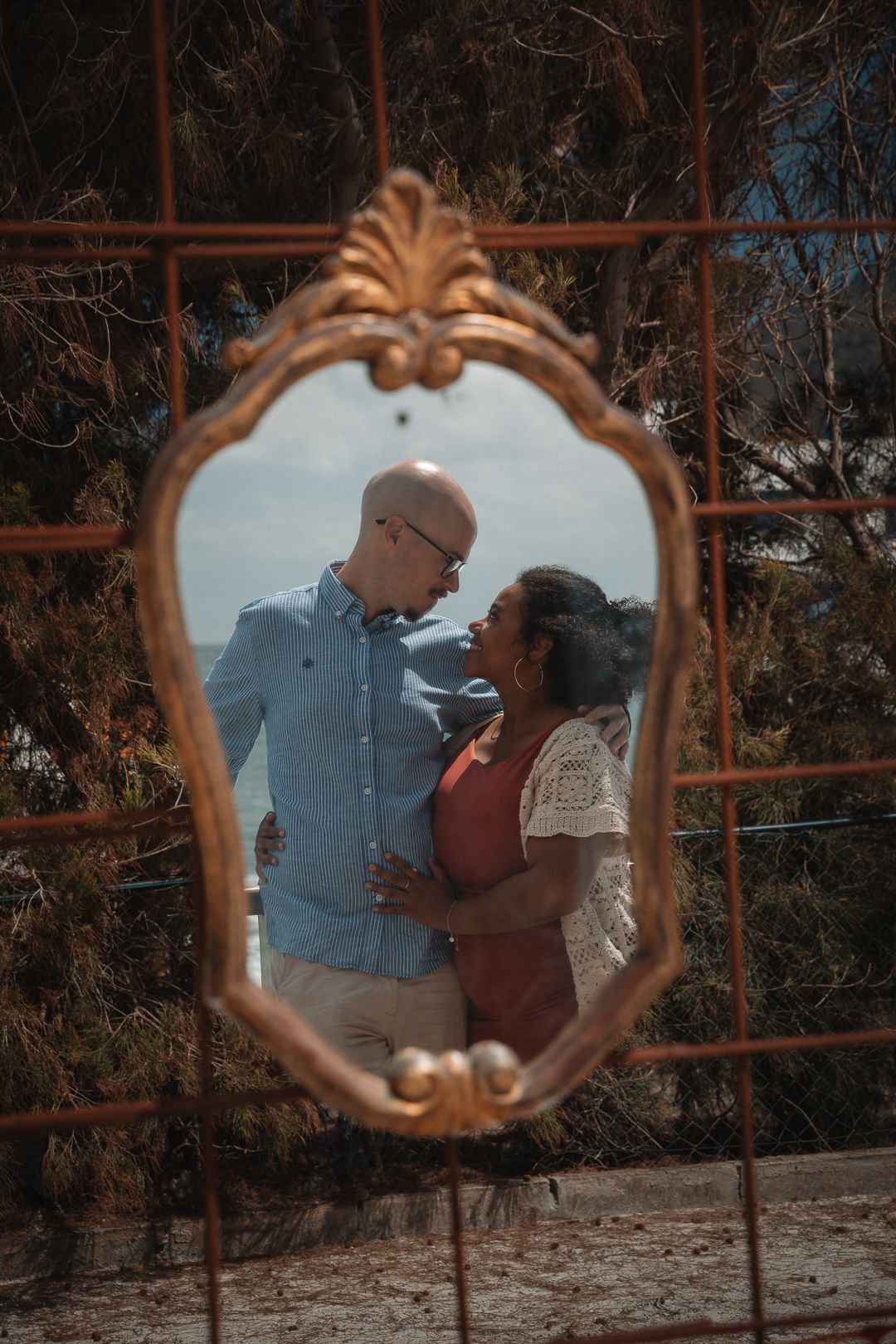 A couple's reflection appears in an ornate vintage mirror frame outdoors.