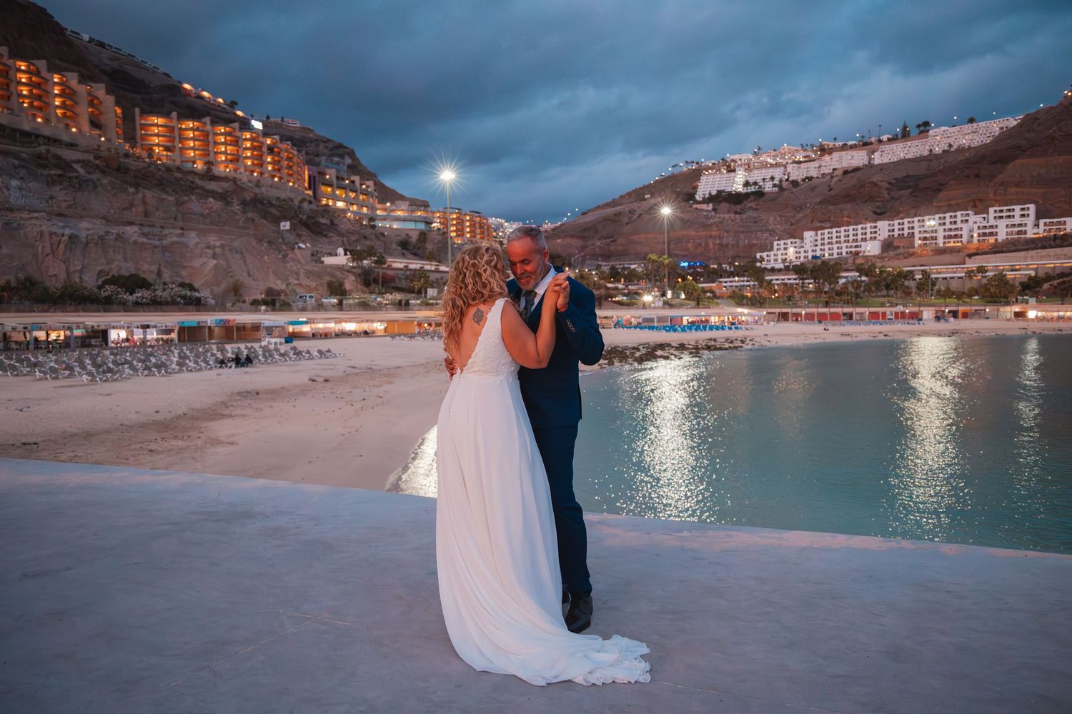 A wedding couple stands on a beach at dusk with dramatic cliffs and illuminated buildings in the background.