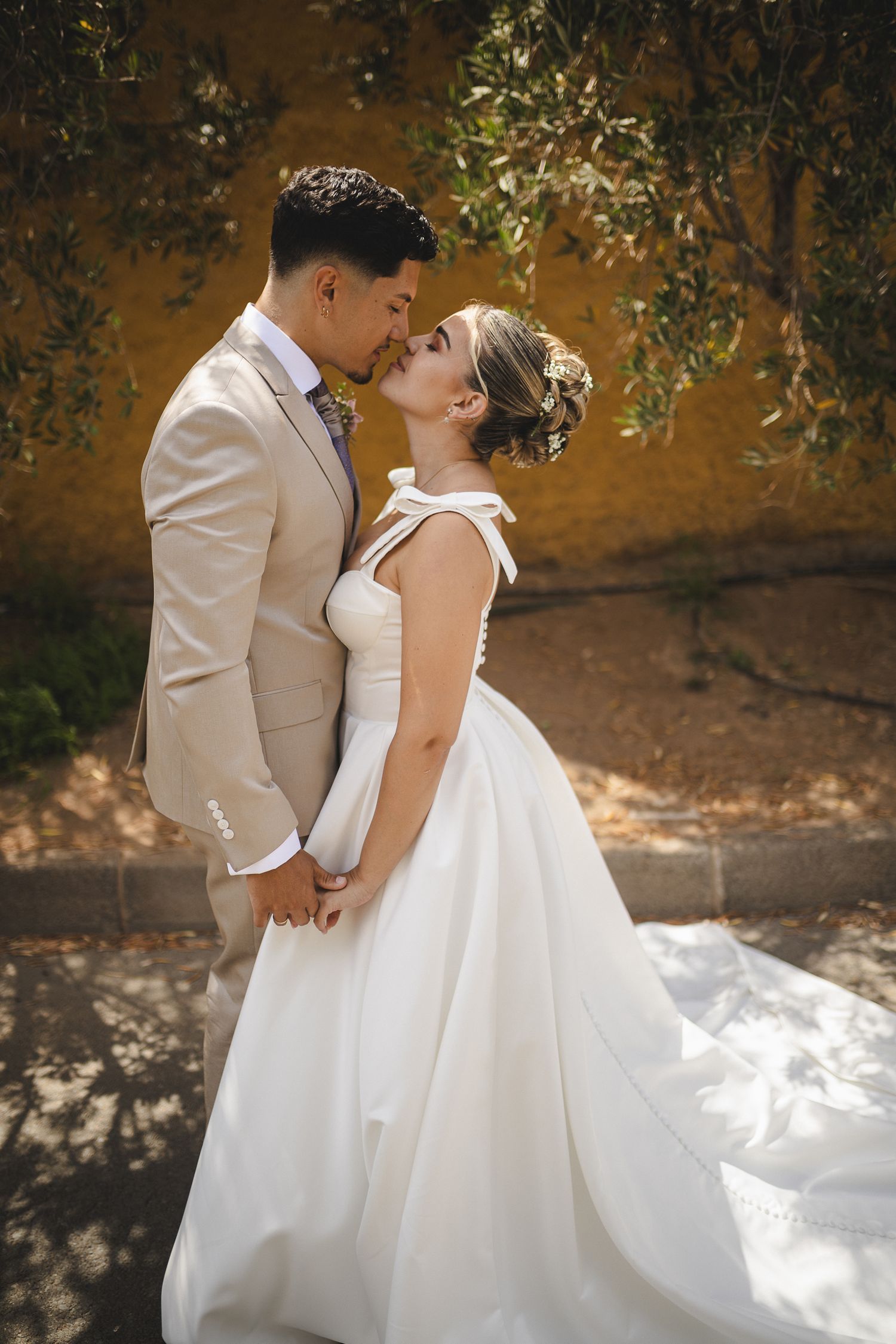 A couple in wedding attire shares a tender moment in dappled sunlight.