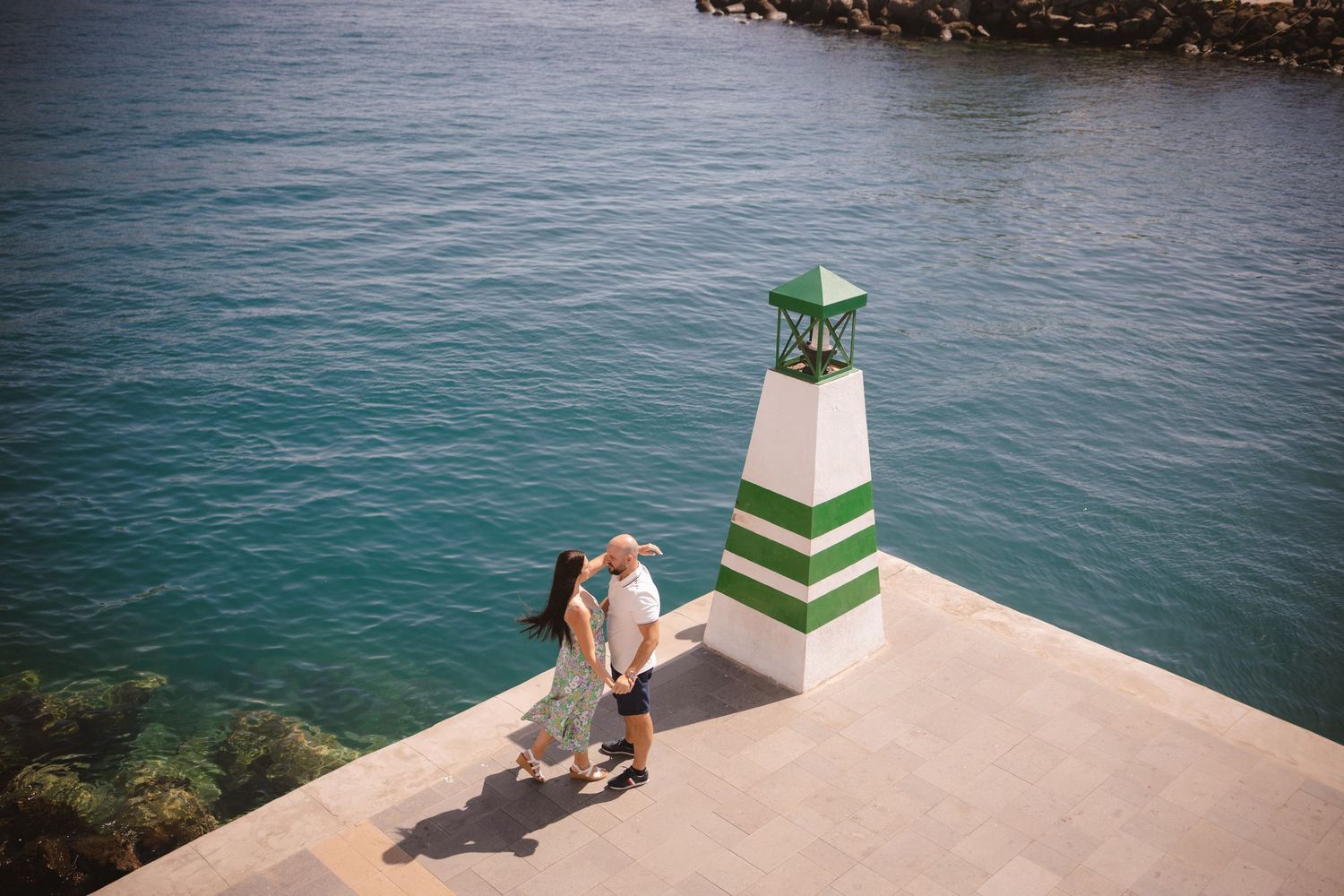 A couple standing on a white lighthouse platform overlooking blue ocean waters.