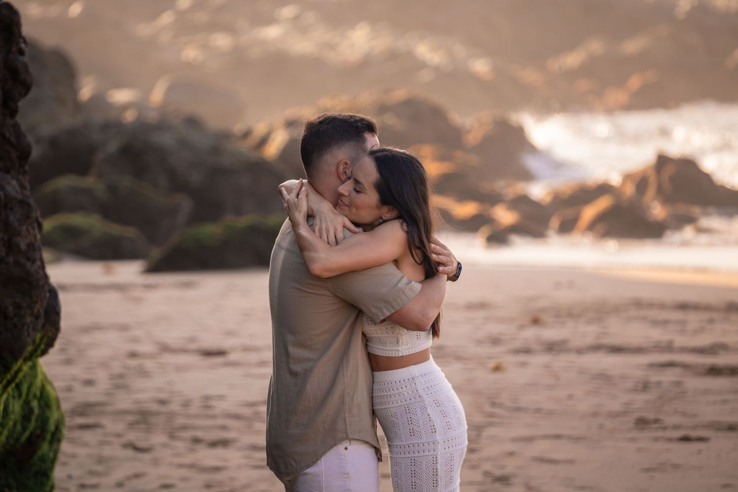 A couple embracing on a beach during golden hour with rocky cliffs in the background.