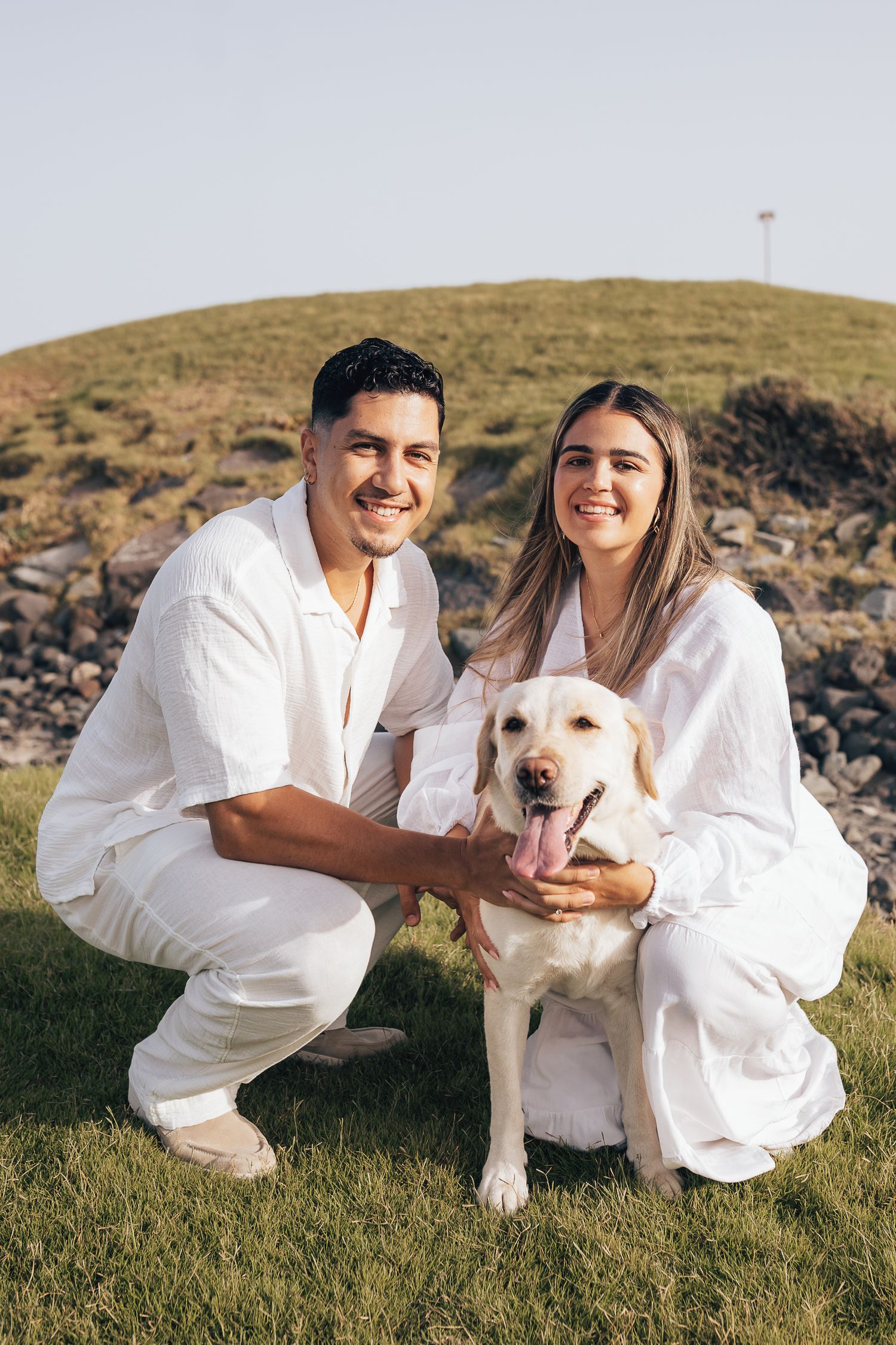 A couple in white outfits poses with their white Labrador dog on a grassy hillside during a photo session.