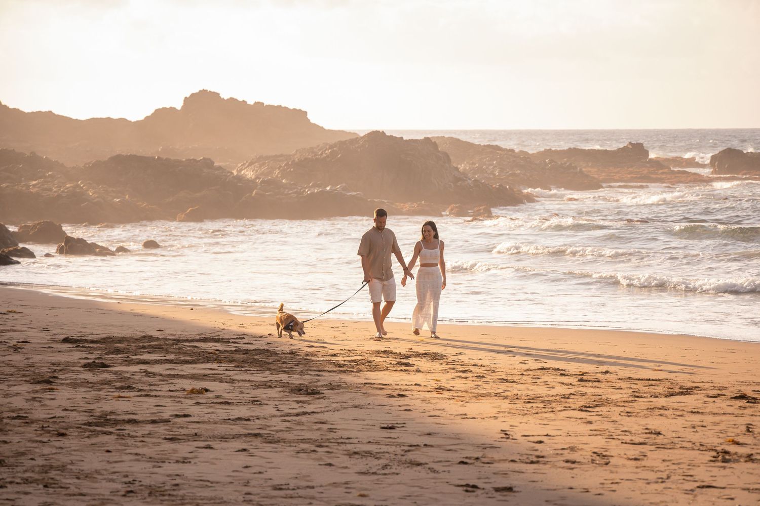 Silhouettes walking along a scenic beach at sunset with rocky coastline in view.