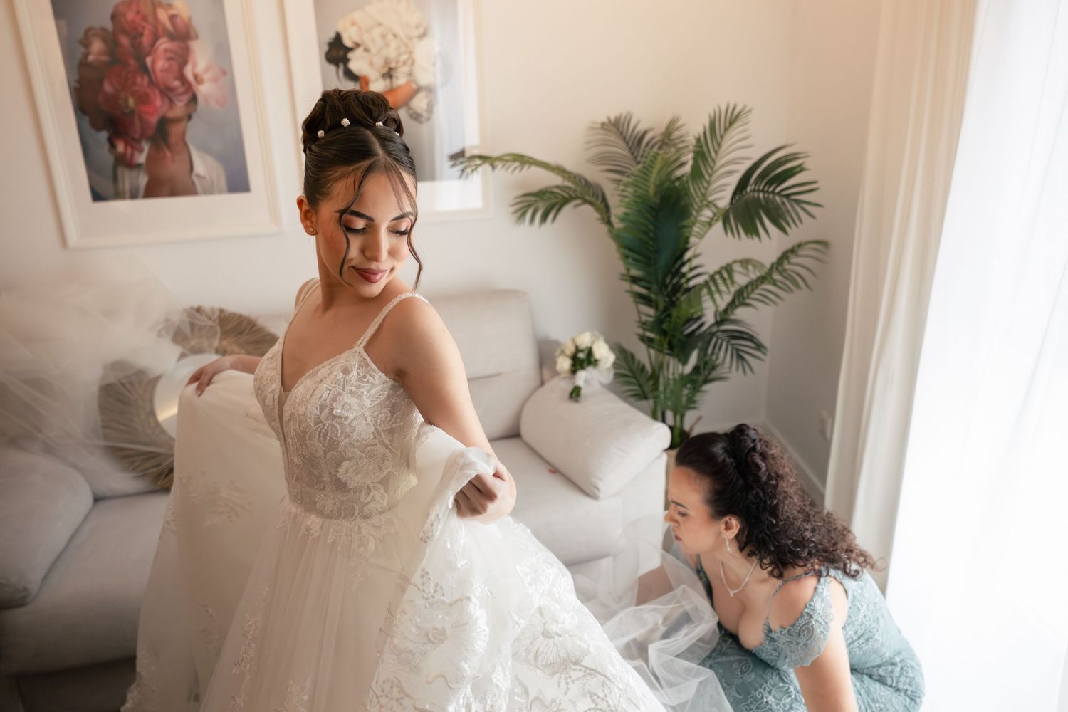 Bride in lace wedding gown being helped by bridesmaid in blue dress in elegant white room with tropical plant.