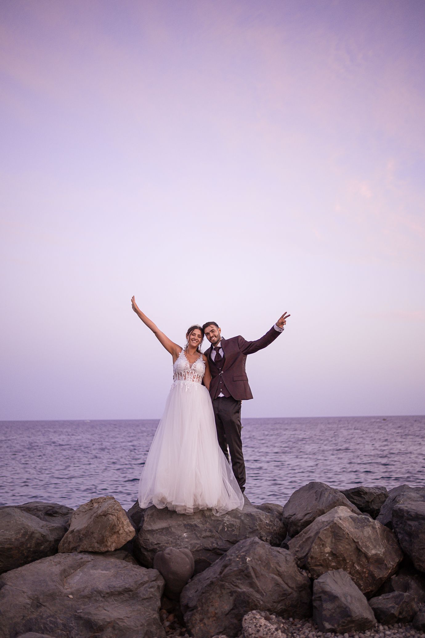 Couple in wedding attire strikes joyful poses on rocky beach at sunset.