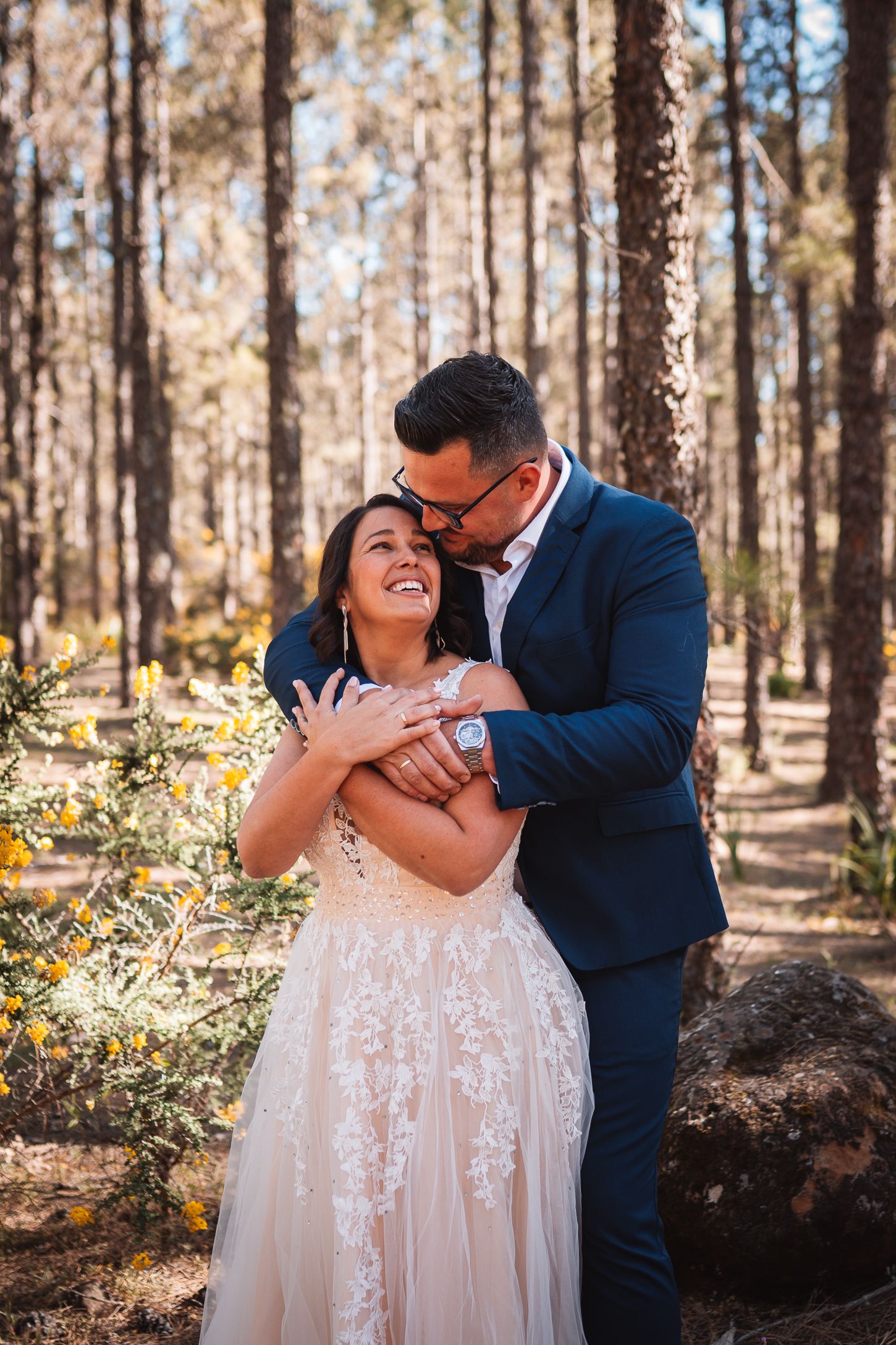 A romantic wedding couple shares a joyful moment together in a sun-dappled forest.