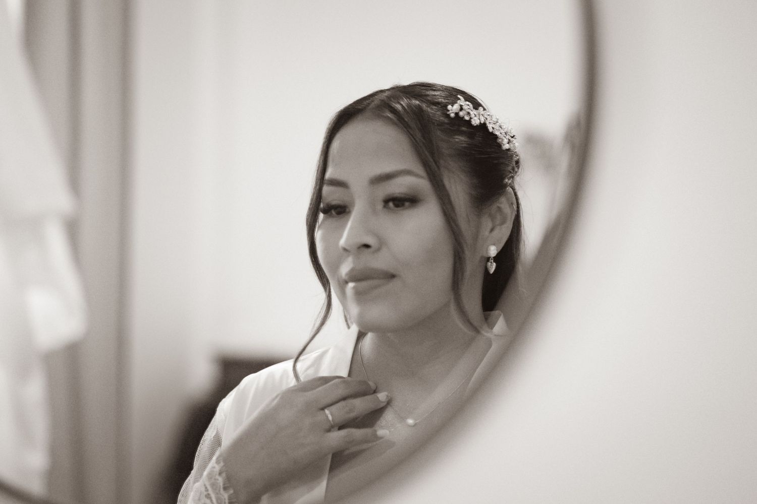 Black and white photo of a bride adjusting her hair, wearing a floral headpiece and elegant earrings, reflected in a mirror.