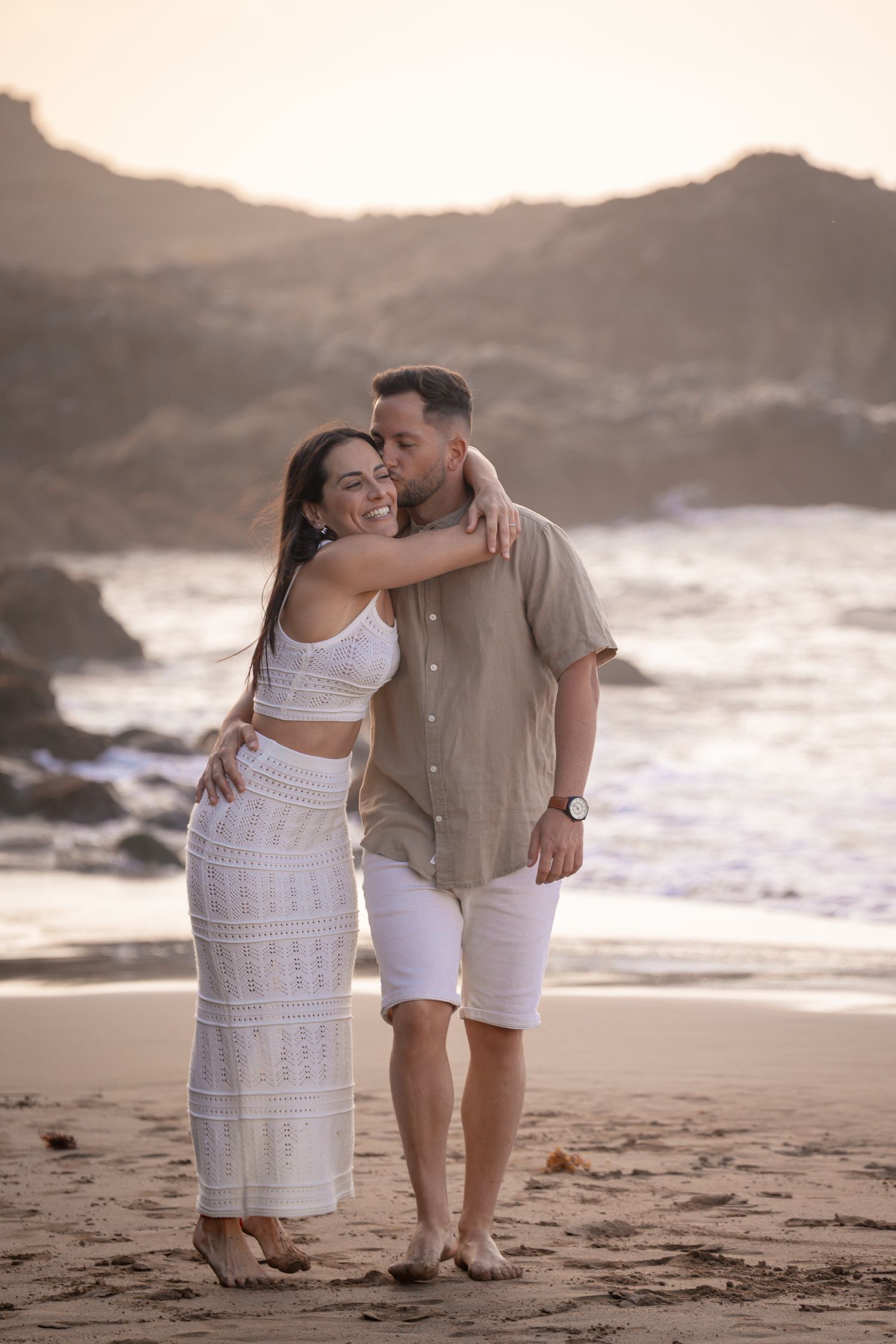 Couple in white outfits sharing tender moment on beach at sunset.
