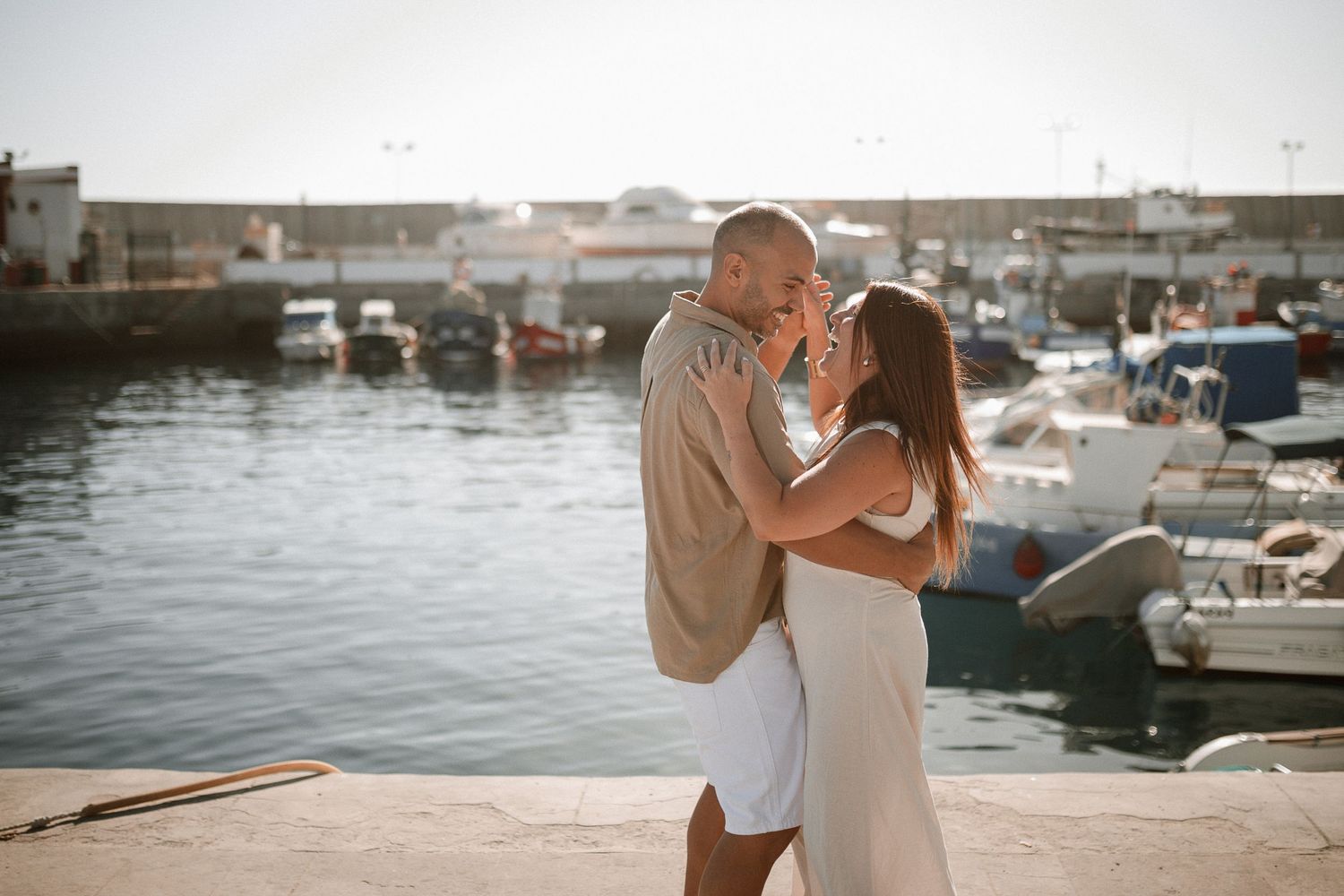 Romantic couple embraces at a marina during sunset with boats and water in the background.