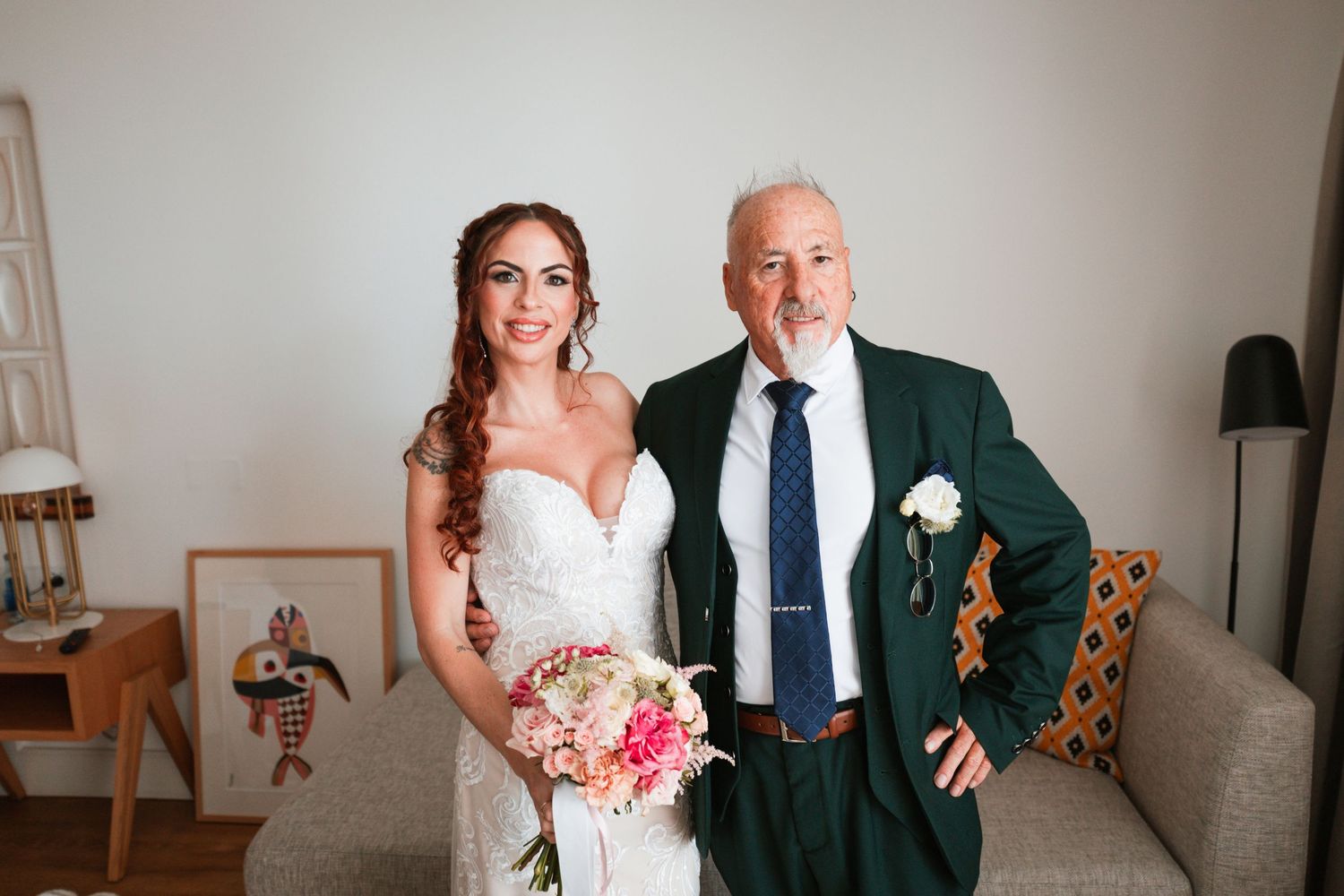 Bride in white lace gown holding pink bouquet poses with older man in green suit at indoor wedding celebration.