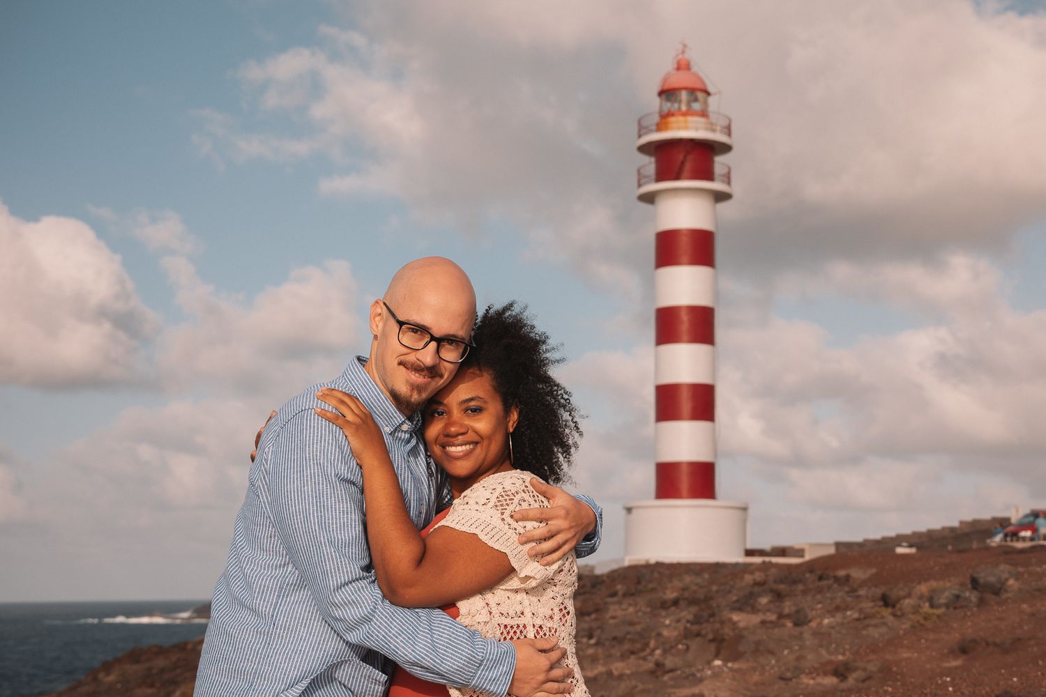 A couple embraces near a red and white striped lighthouse under cloudy skies.