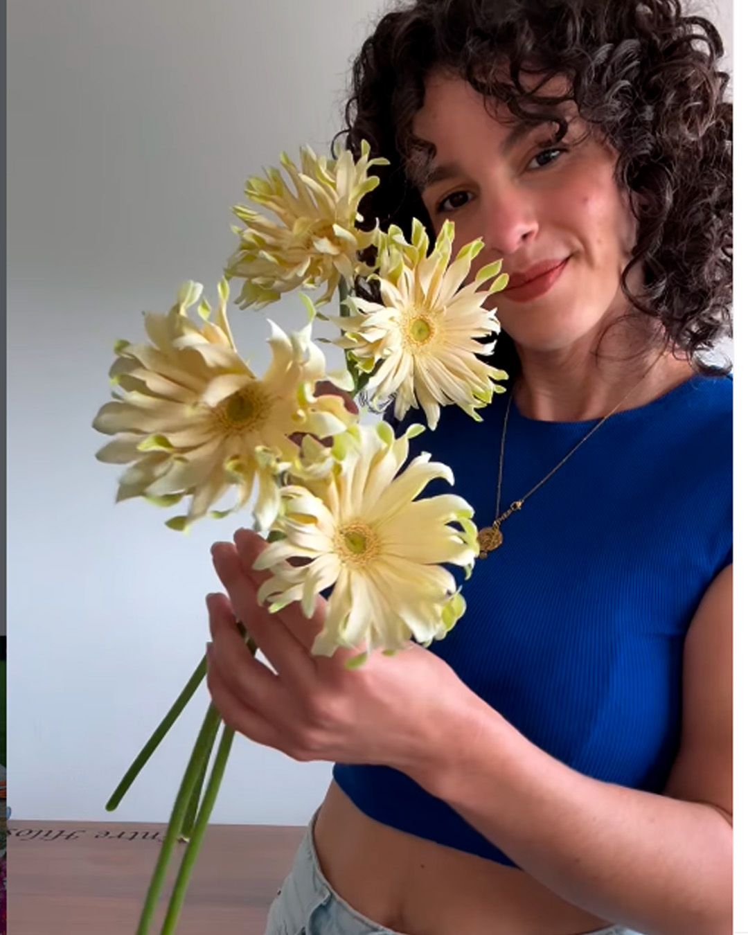 Someone in a blue crop top holds pale yellow dahlia flowers against a light background.