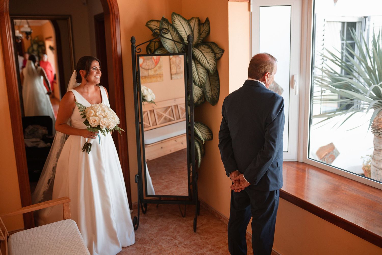 A bride in white dress stands near a mirror while someone waits in the background.