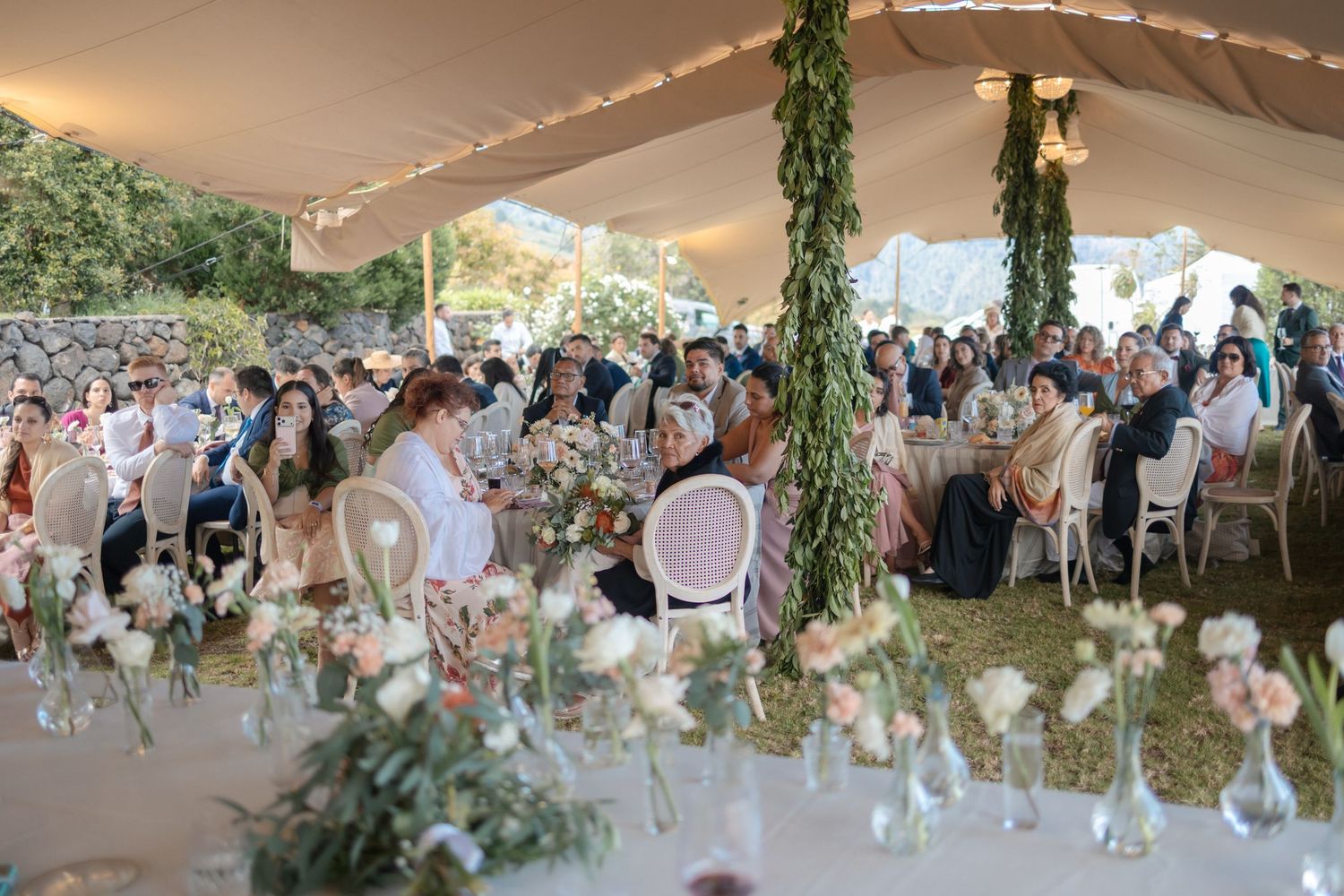 Elegant outdoor wedding reception under a tent with floral decorations, guests seated at tables with pink and white flowers.