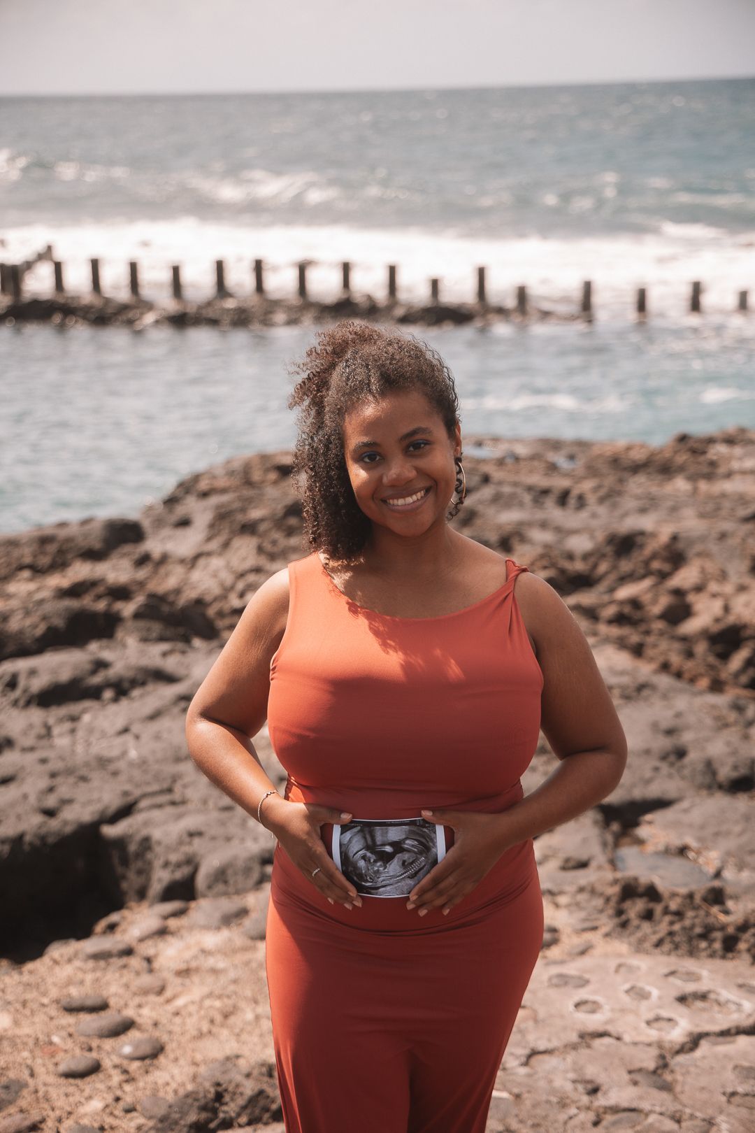 Couple poses for romantic maternity photos on a rocky beach with ocean waves in the background.