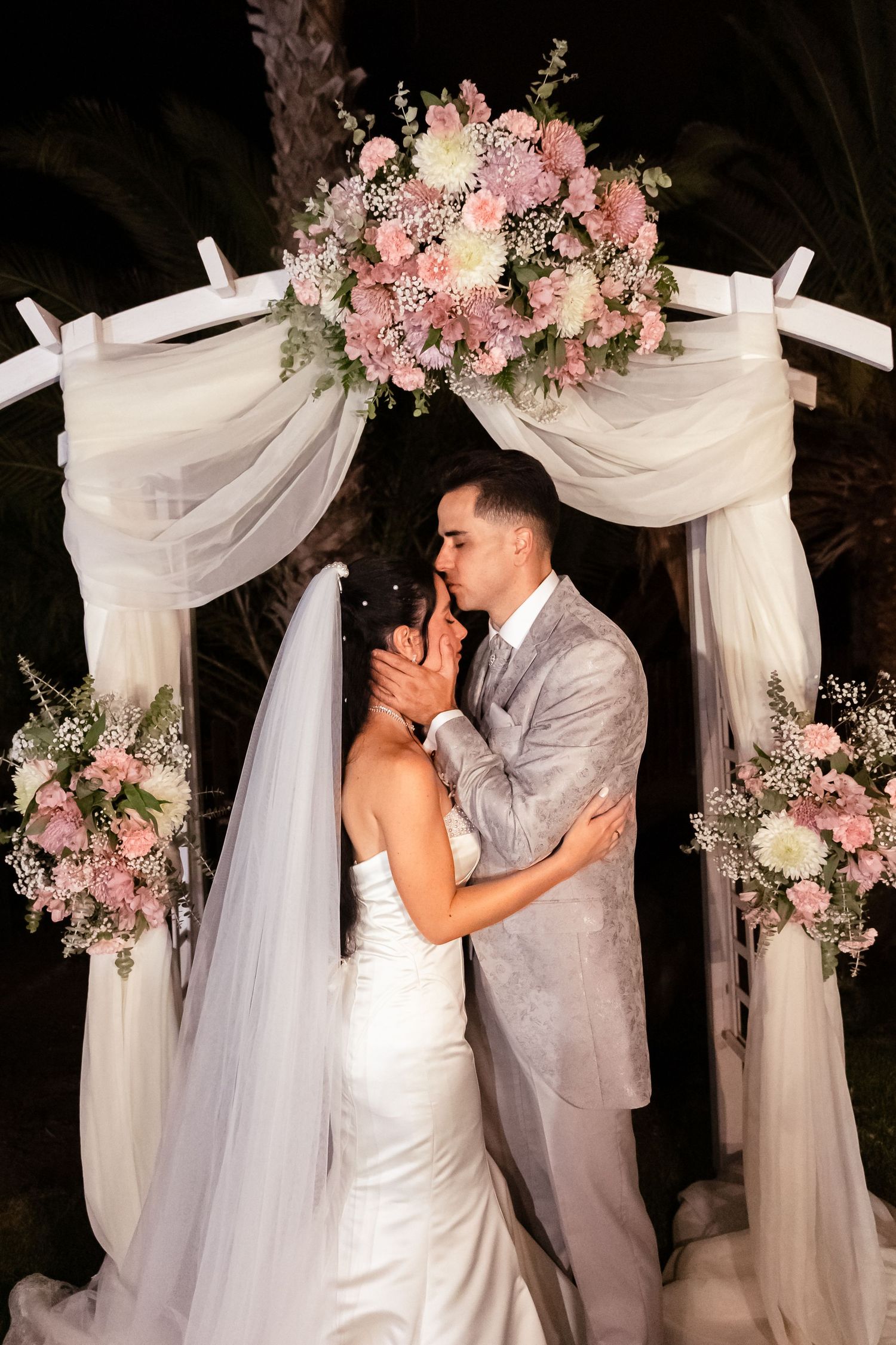 A couple shares an intimate moment under a floral arch decorated with pink blooms and white fabric.