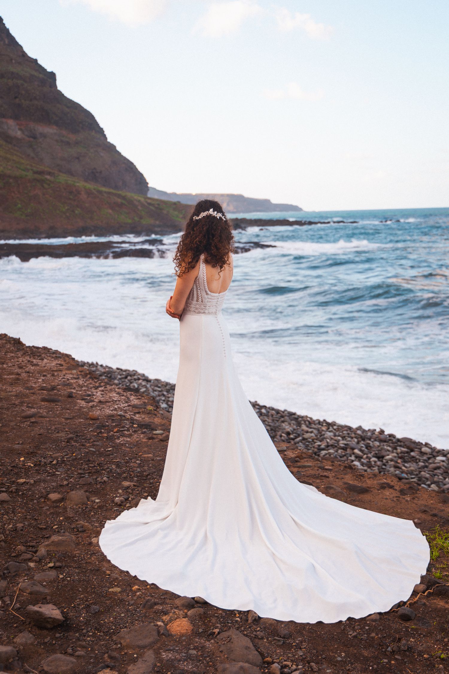 A flowing white wedding dress with a long train cascades over rocky coastal terrain as waves crash against the shore.