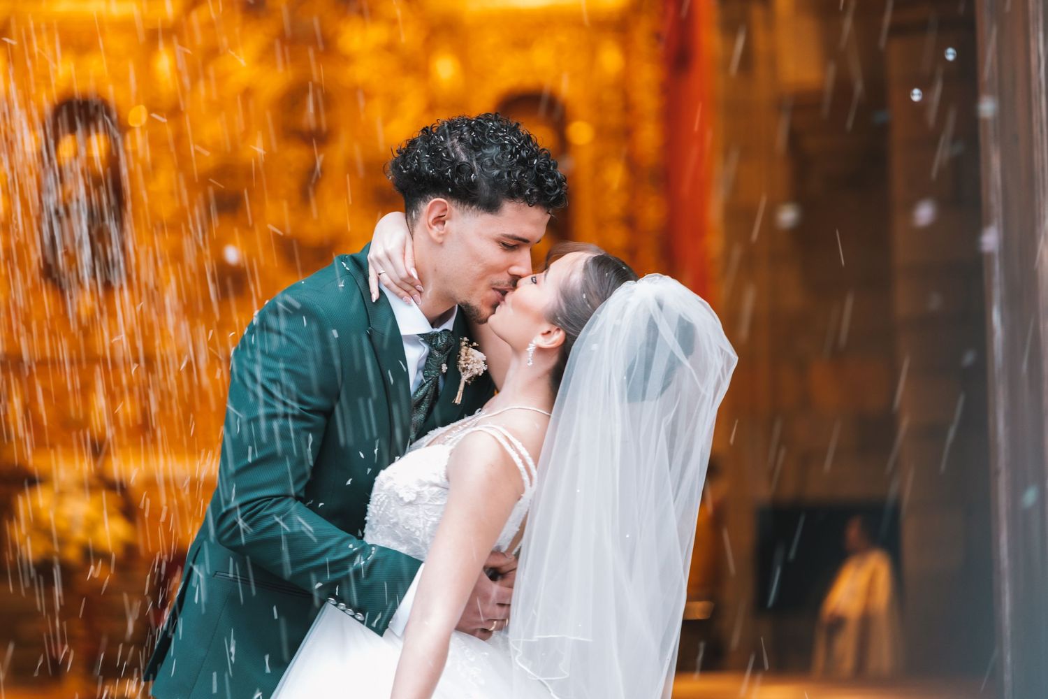 A couple in wedding attire share a romantic moment in front of a glowing orange background with falling rain.