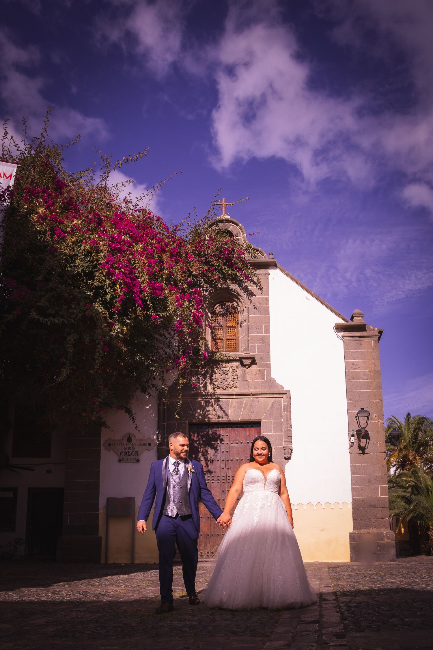 Wedding scene against white colonial architecture with vibrant purple sky and bougainvillea.