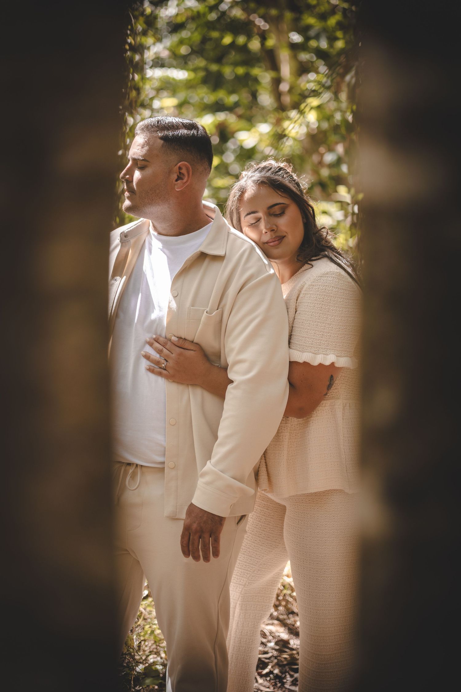 Couple in matching beige outfits embracing outdoors in dappled sunlight among trees.