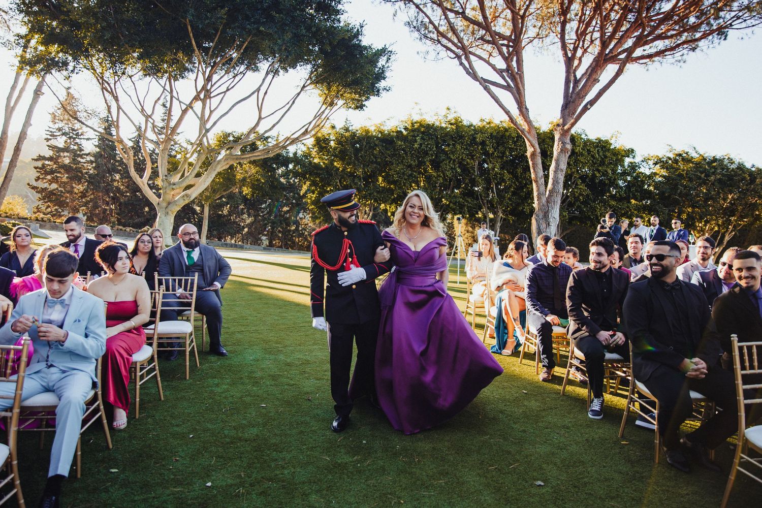 Outdoor wedding reception setup with guests seated at long tables under trees.