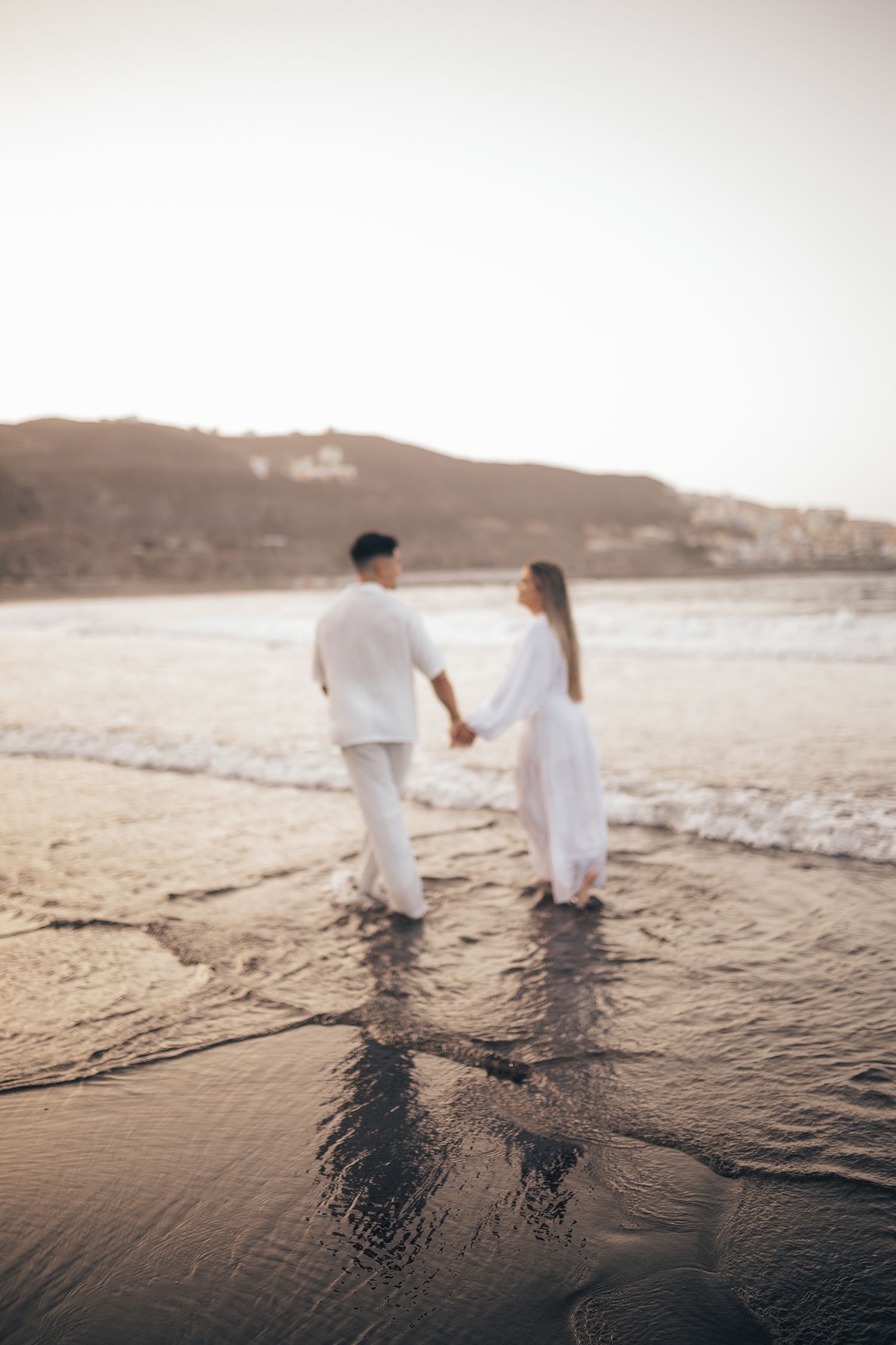 Two people in white clothing walk along a beach shoreline at sunset creating silhouettes.