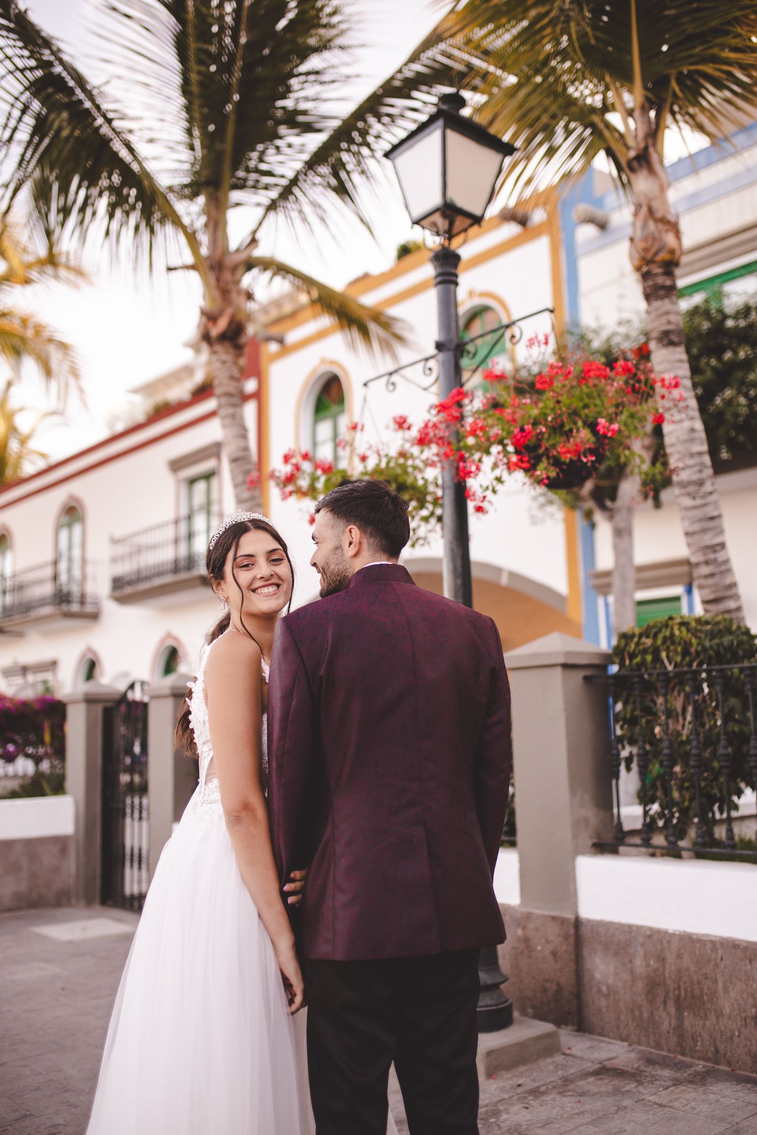 A romantic wedding couple stands on a charming street with palm trees and Spanish colonial architecture in the background.