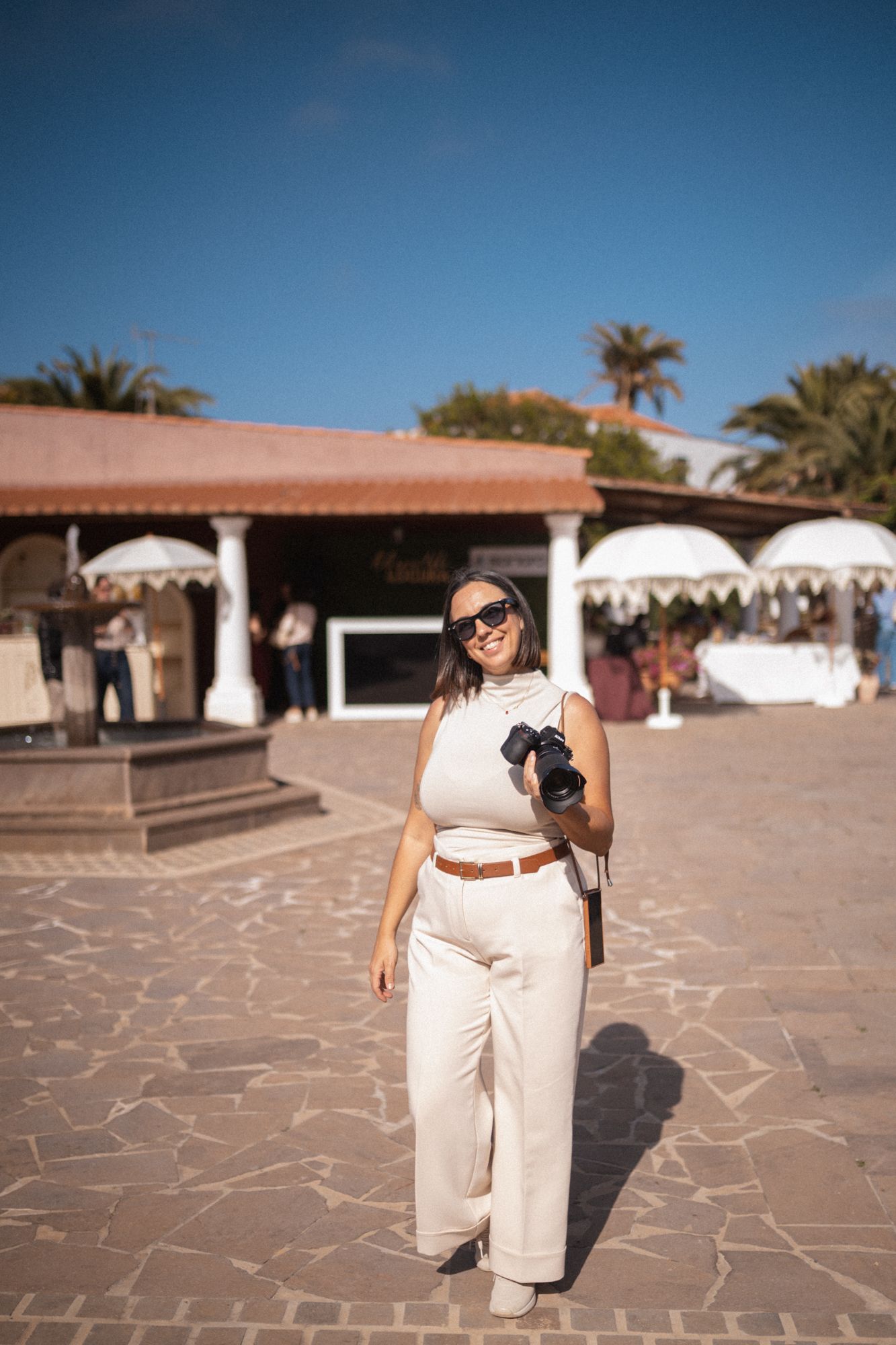 A person in a white outfit and sunglasses poses in a Mediterranean-style courtyard with stone paving and palm trees.