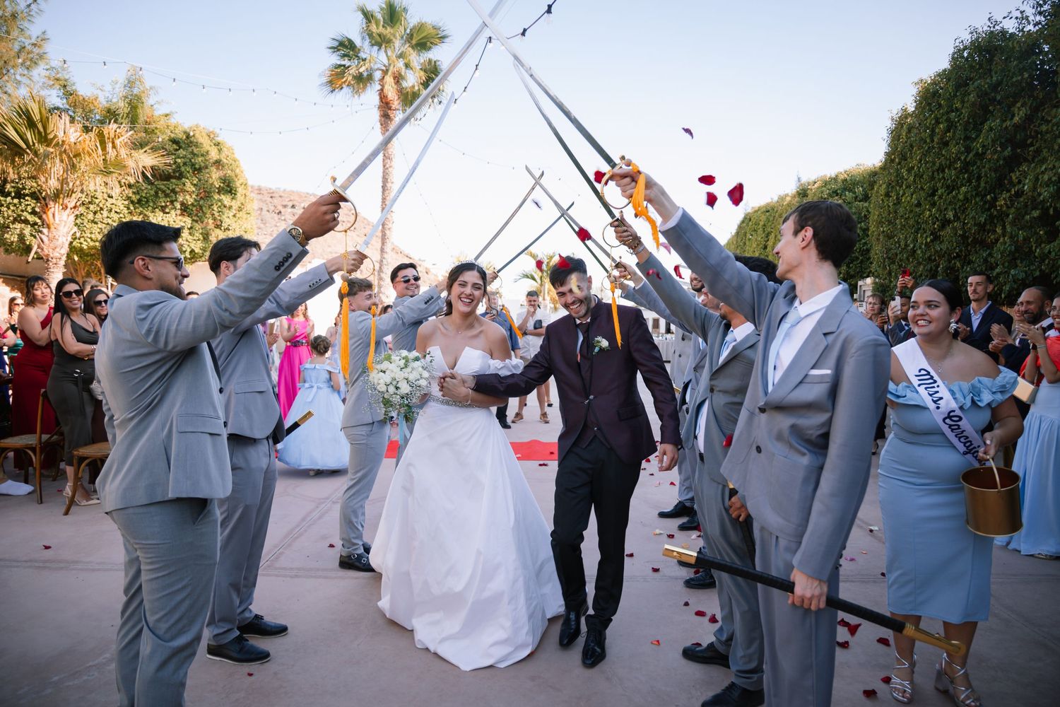 Wedding party members in gray suits and white dresses dancing outdoors near palm trees.