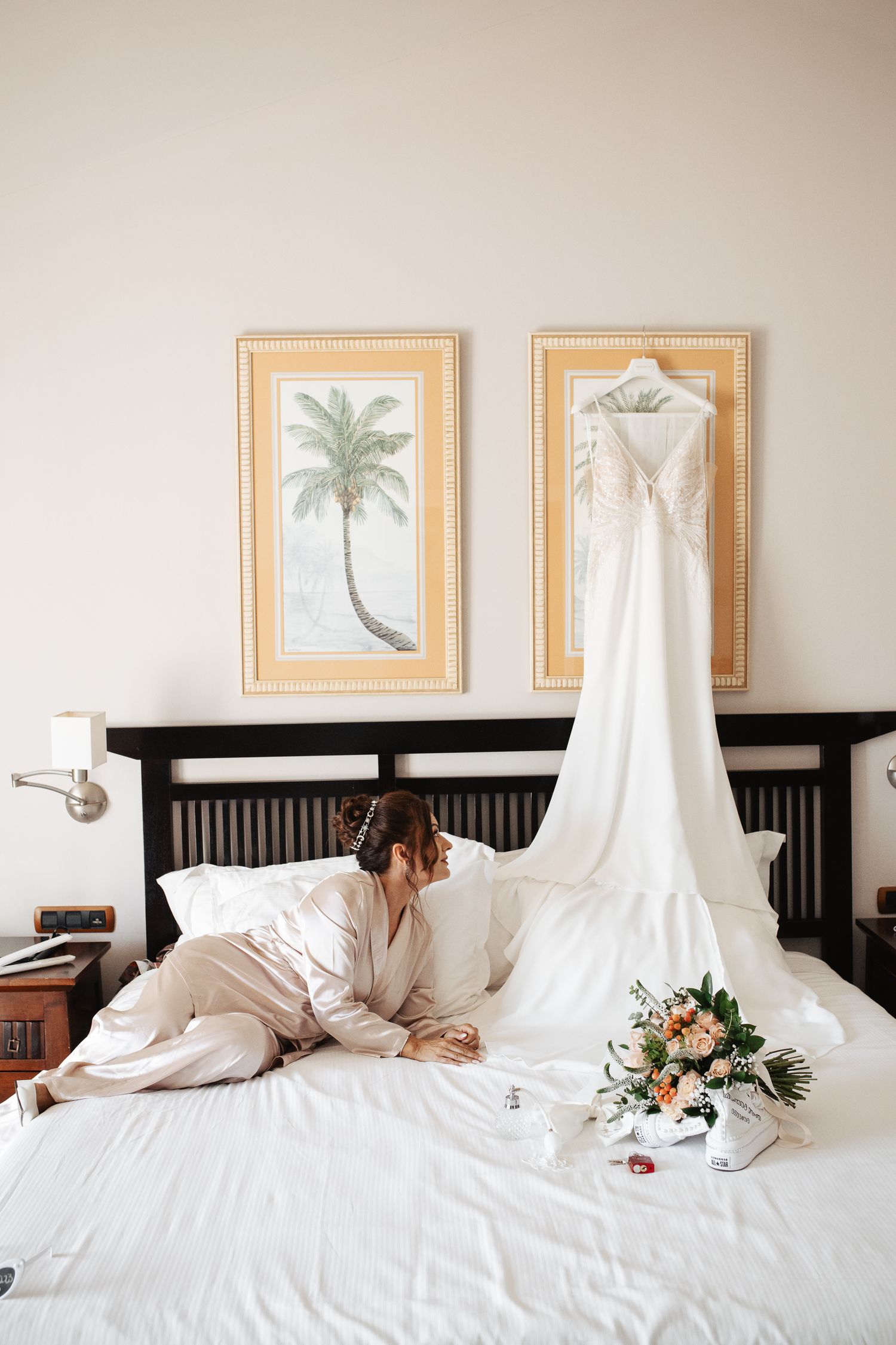 Wedding dress draped across white bed with palm tree artwork and black headboard.