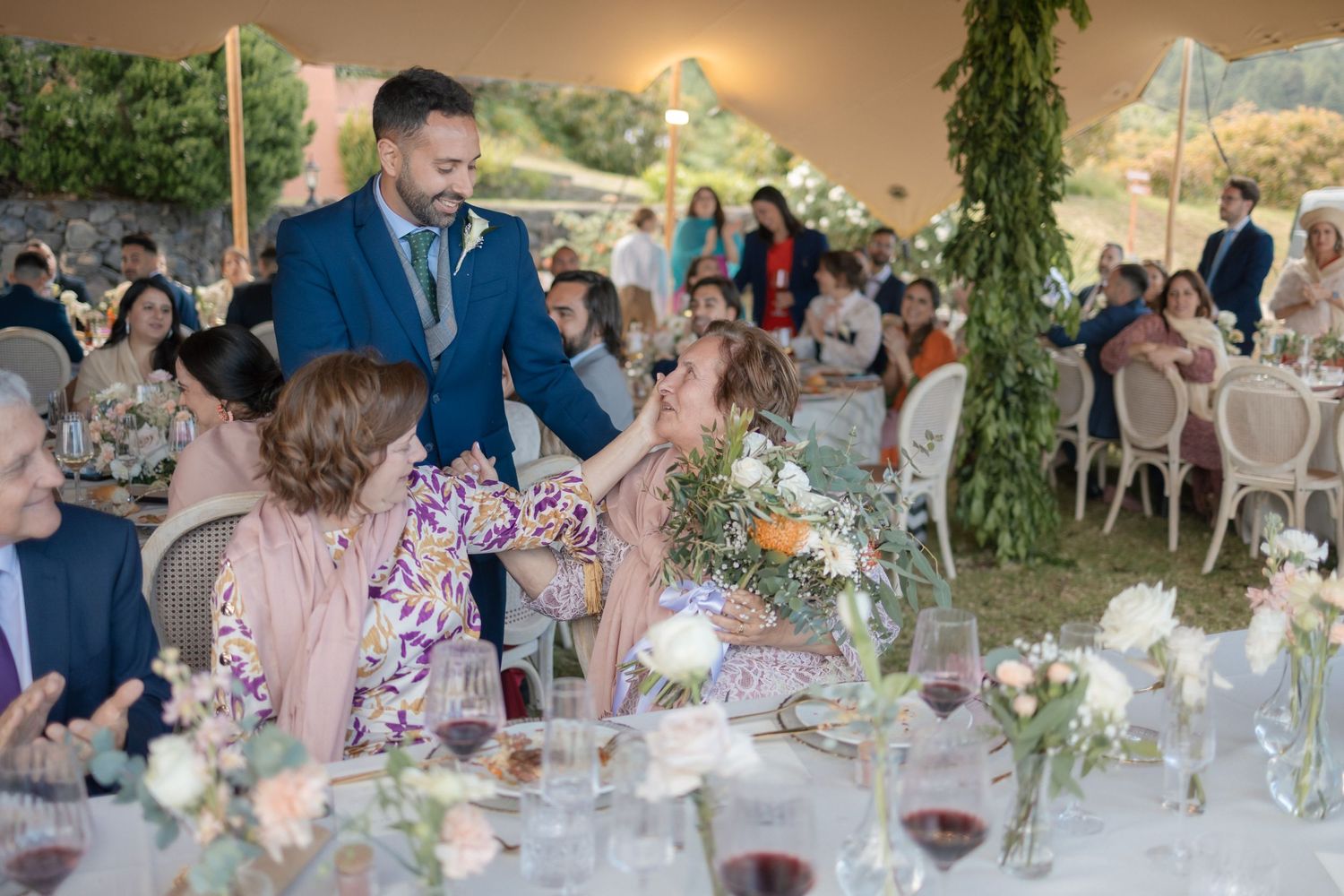 Groom in blue suit greets seated wedding guests at elegant outdoor reception with floral centerpieces.