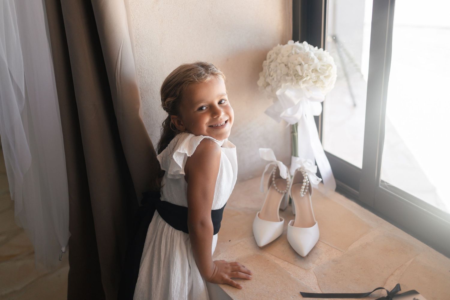 Smiling flower girl in white dress with navy sash stands by window near bridal bouquet and white heels.