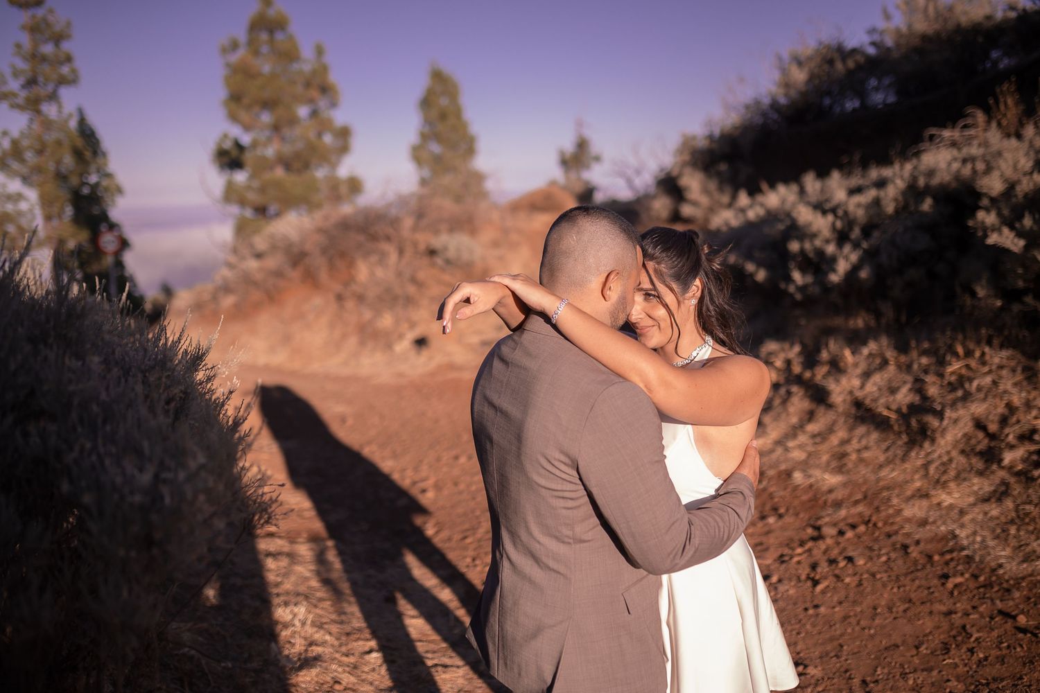 A couple embraces on a dirt path during sunset, casting long shadows.
