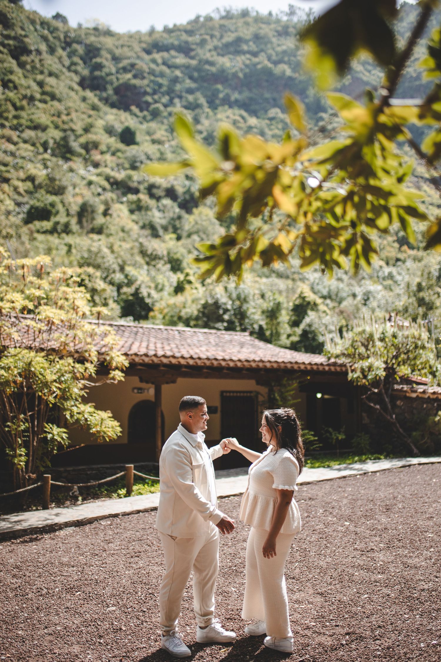 Couple in white attire holding hands outdoors with lush green mountains and a rustic house in the background.