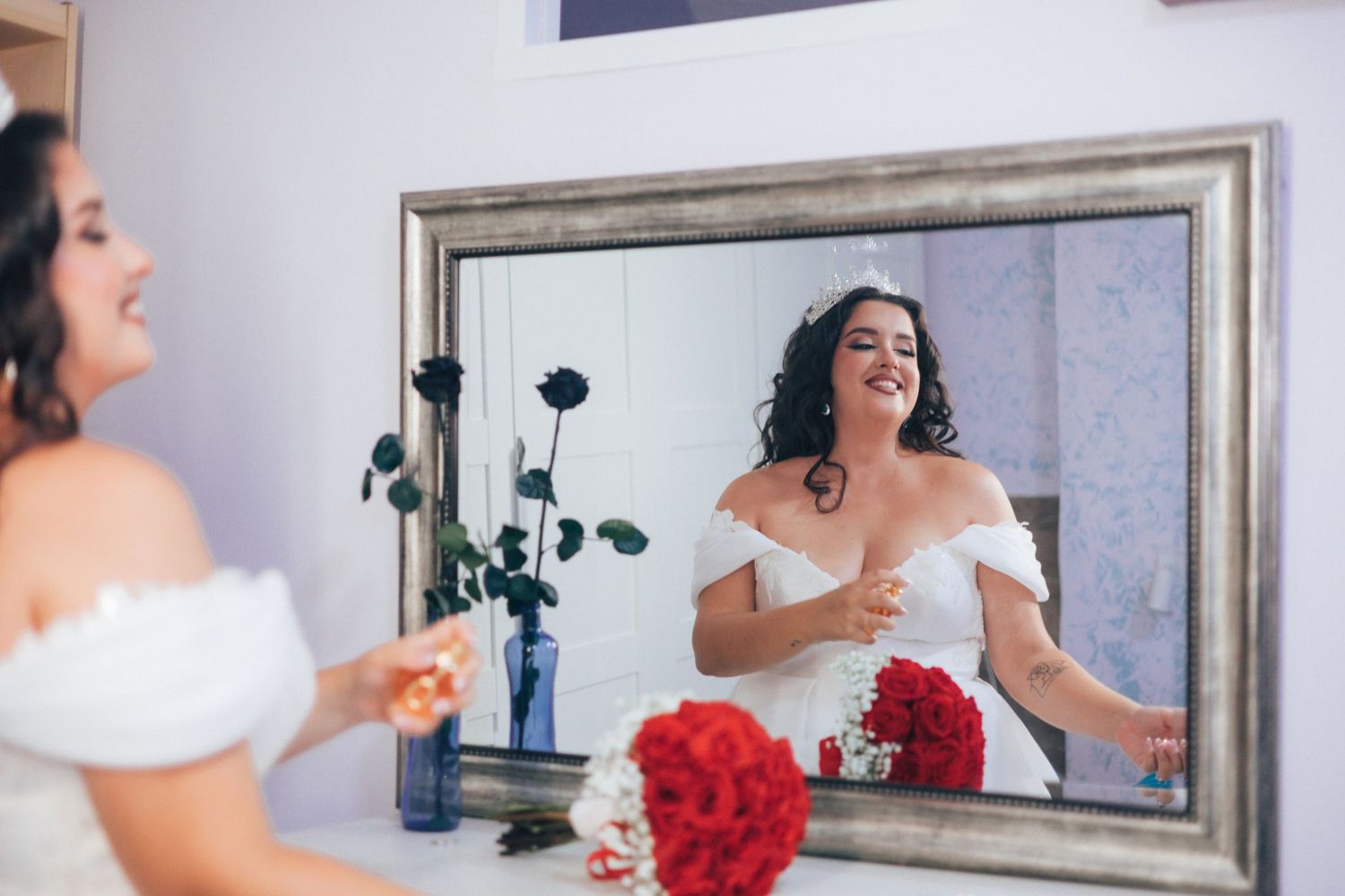 Bride getting ready in front of mirror while holding red bouquet in elegant white wedding dress.