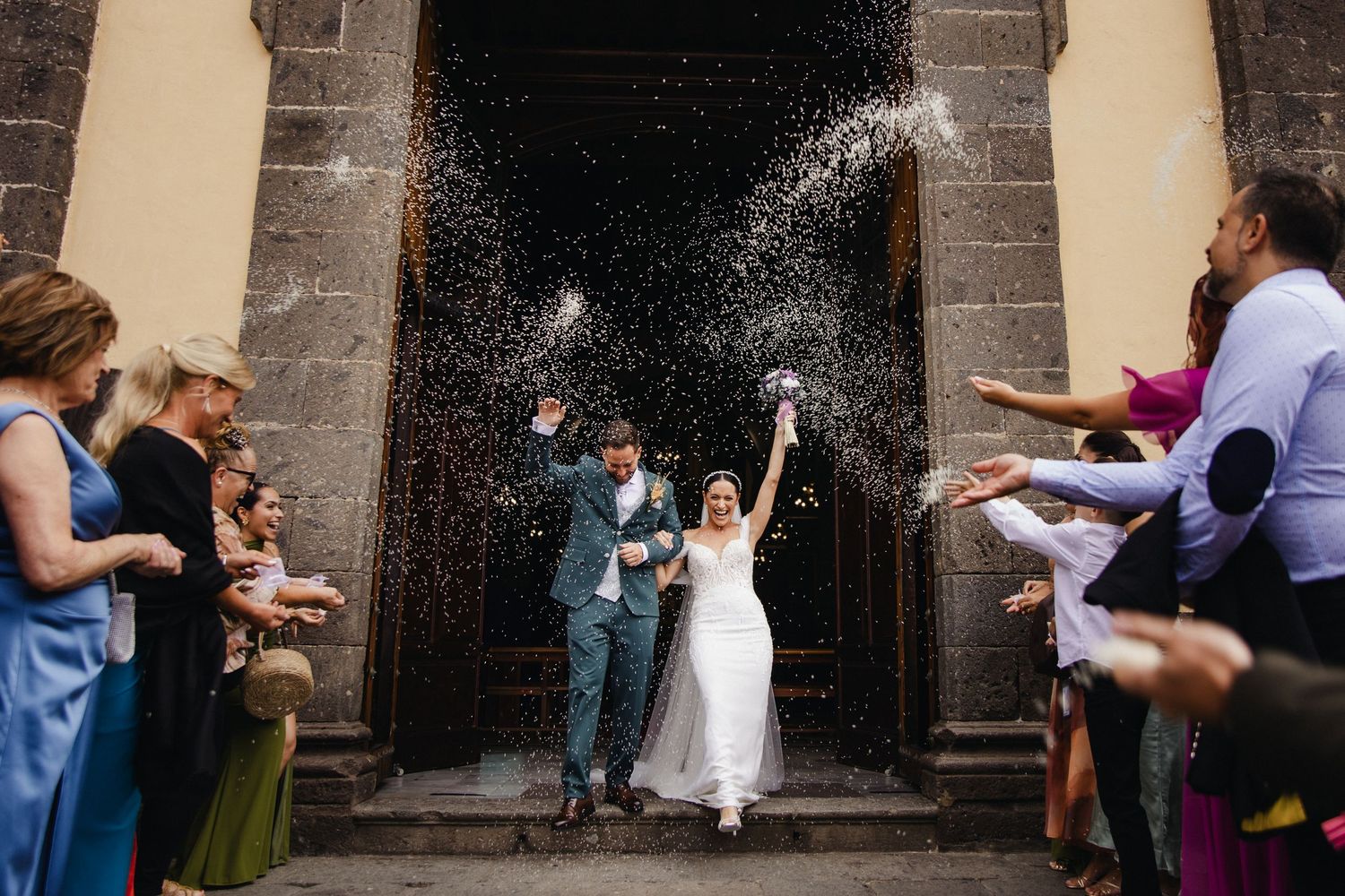 Elegant wedding party posing in front of historic stone building with classic vintage cars.