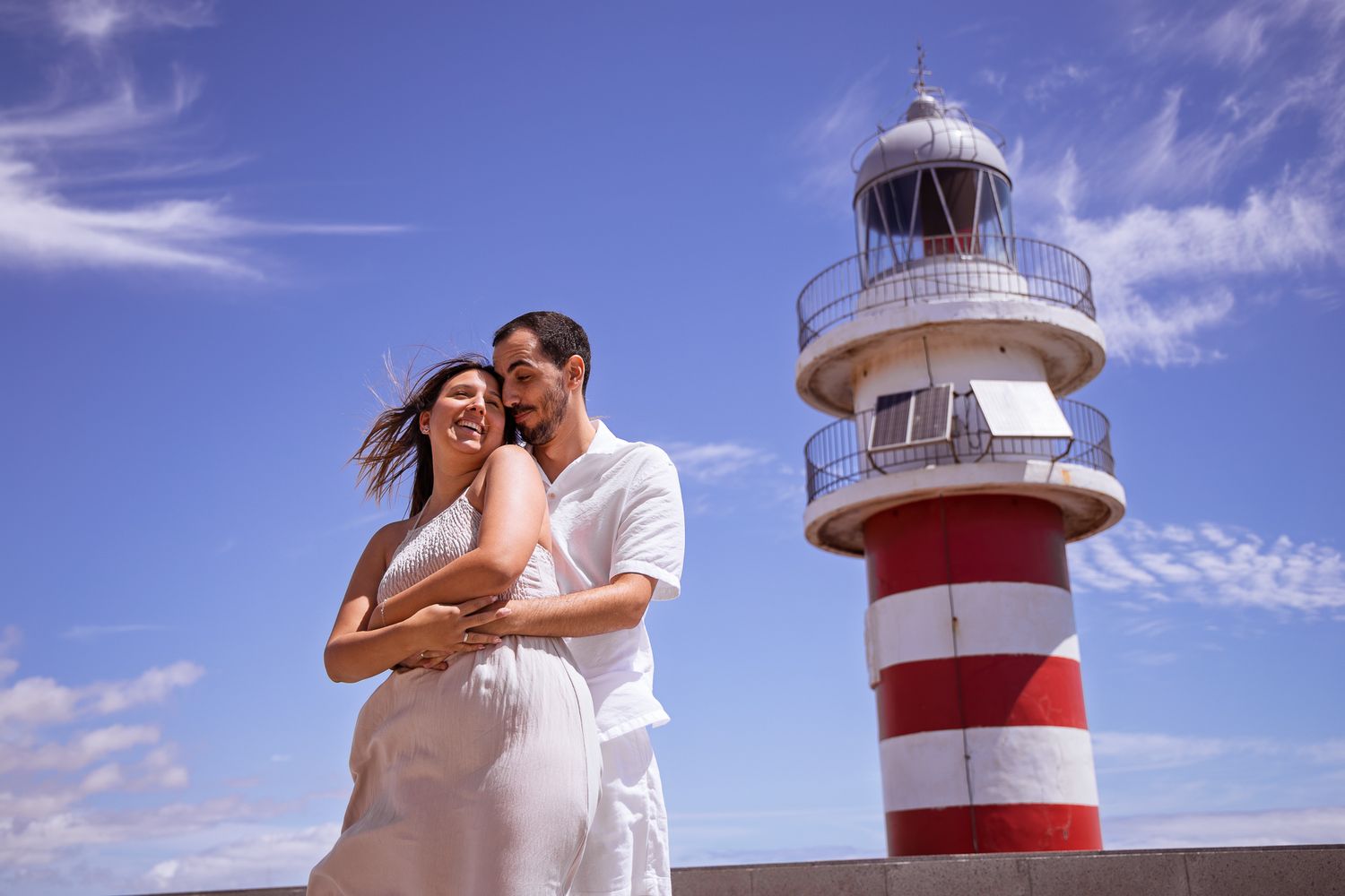 Romantic couple photos by red and white lighthouse against blue sky.