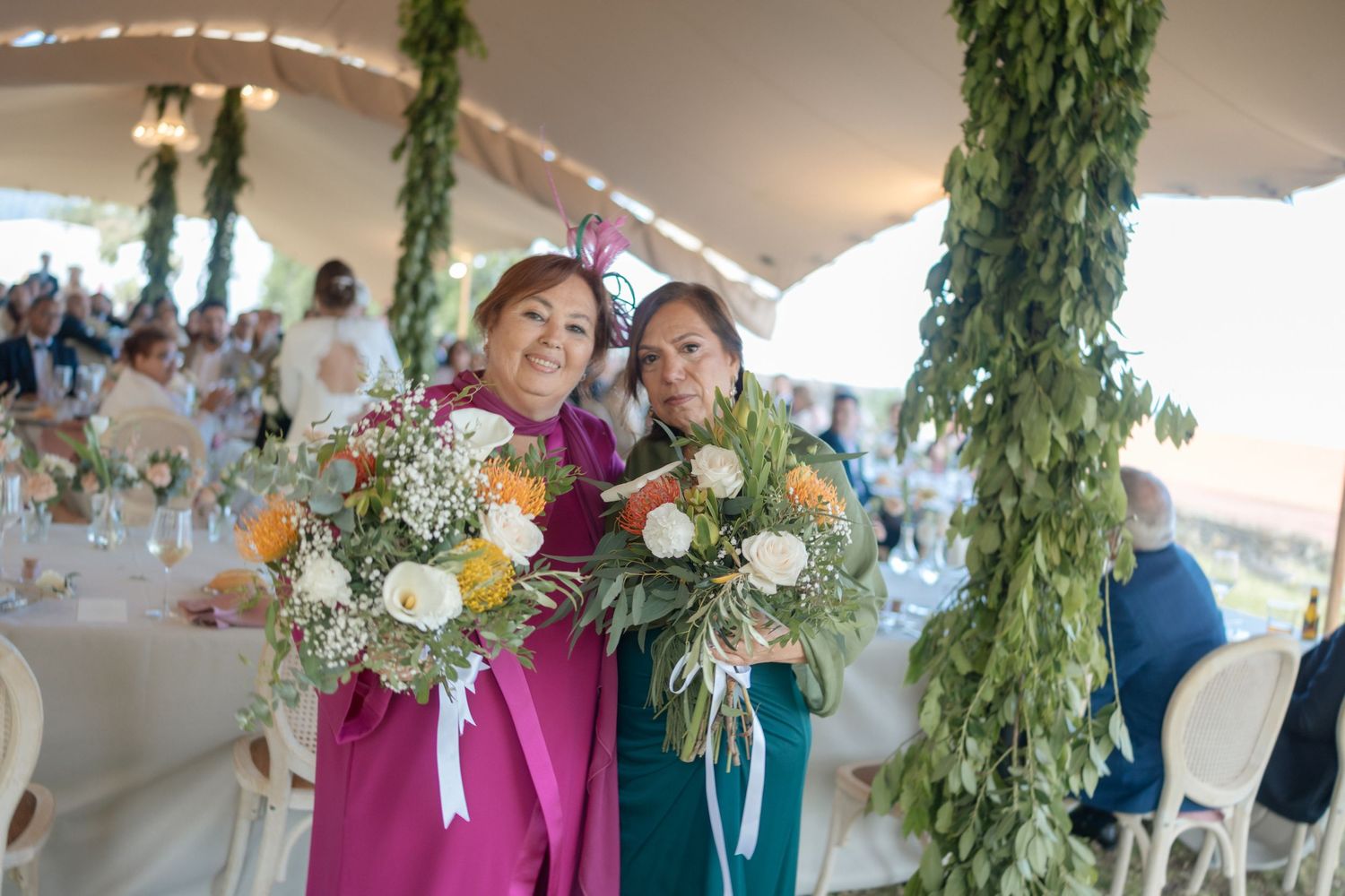 Two women in colorful dresses holding bouquets of flowers at an elegant outdoor wedding reception.
