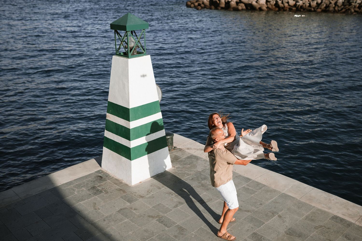 A couple enjoys romantic moments near a striped lighthouse by the ocean.