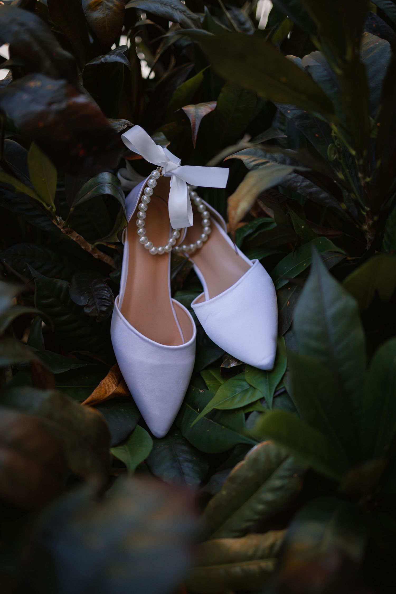 Elegant white bridal shoes with pearl details nestled among dark green leaves.